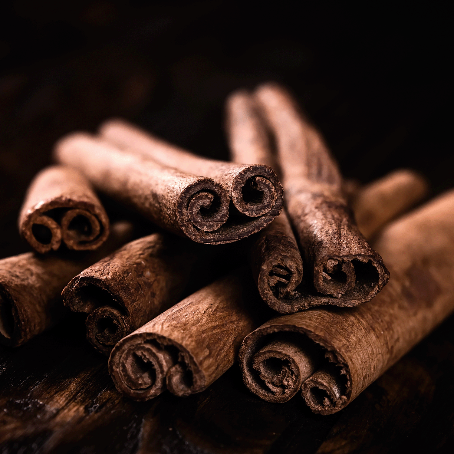 Several cinnamon sticks are stacked on a dark wooden surface, reminiscent of the cozy aroma from Tuscany Candle® SEASONAL’s Grizzly Ghouls: Autumnal Spices & Fogged Musk Scented Halloween Candle (14 oz).