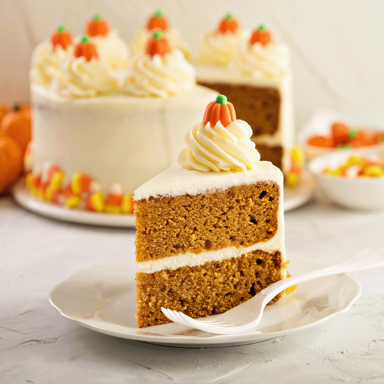 A slice of pumpkin spice cake with white frosting sits beside the Tuscany Candle® SEASONAL Pumpkin Cake Pops: Spice Cake & Vanilla Icing Scented Halloween Candle (18 oz), with candy corn and festive decorations in the background.