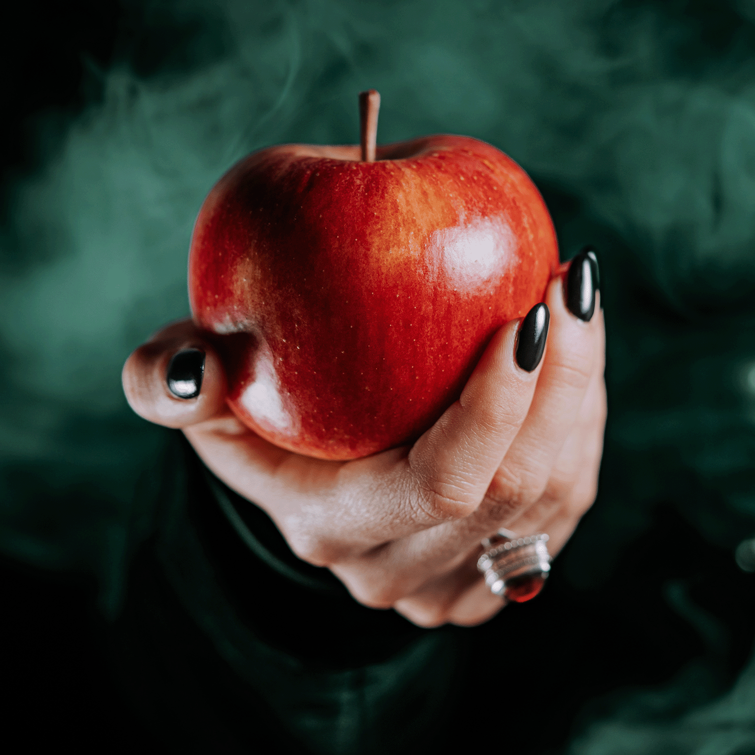 A hand with black nails and a silver ring holds a shiny red apple against a dark background—evoking the allure of Tuscany Candle® SEASONAL's Enchanted Apple: Caramel & Poisoned Apple Scented Halloween Wax Melt (2.5 oz).