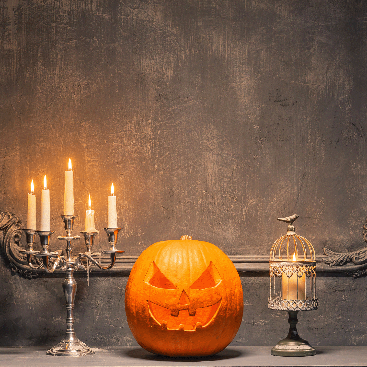 A Charmed Pumpkin: Black Cardamom & Pumpkin Scented Halloween Candle (12 oz) from the Tuscany Candle® SEASONAL Spooky Skull Collection sits with a jack-o'-lantern, candelabra, and birdcage for cozy autumn decor against a gray textured wall.