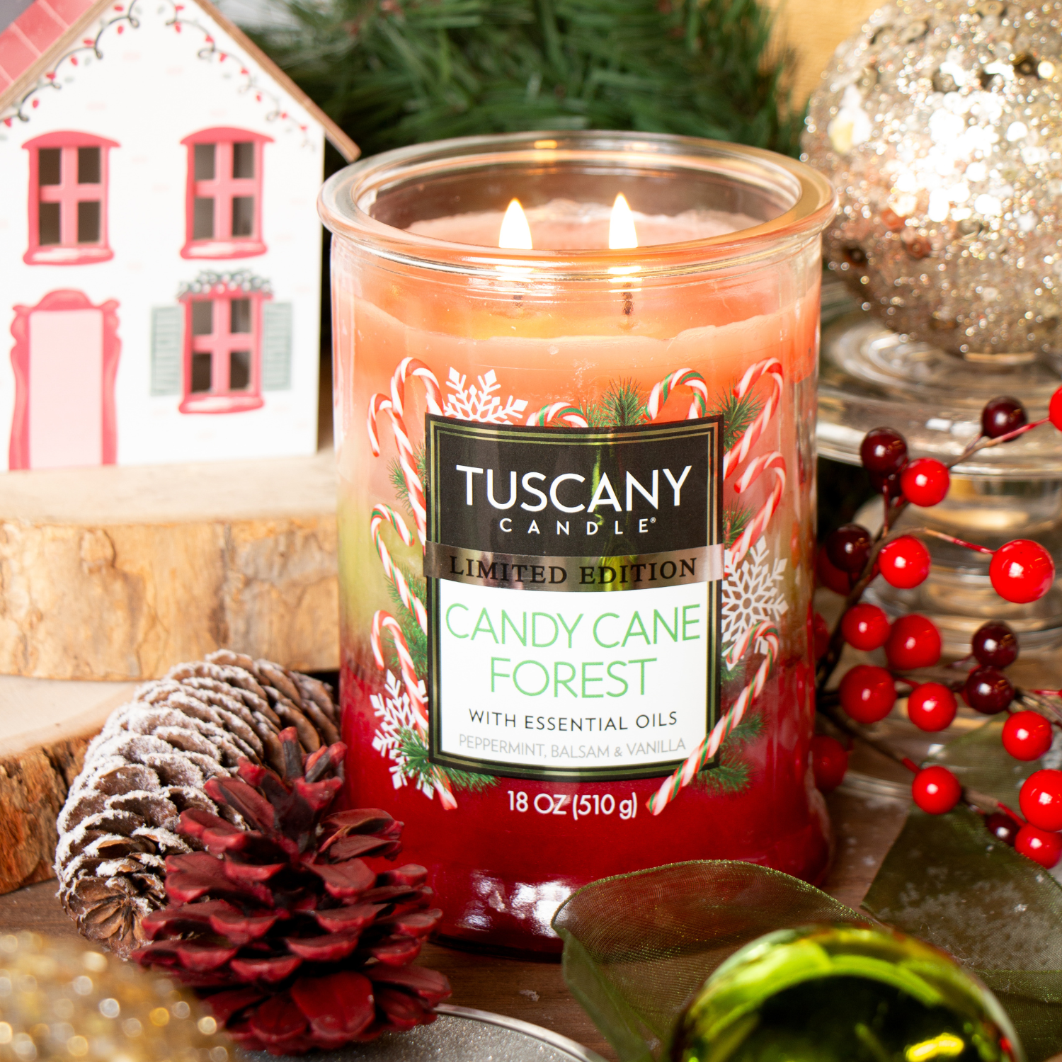 The Tuscany Candle® SEASONAL "Candy Cane Forest: Peppermint & Balsam Scented Holiday Candle" (18 oz) is displayed among pinecones, berries, a cozy house, and festive greenery, creating a cheerful holiday vibe.