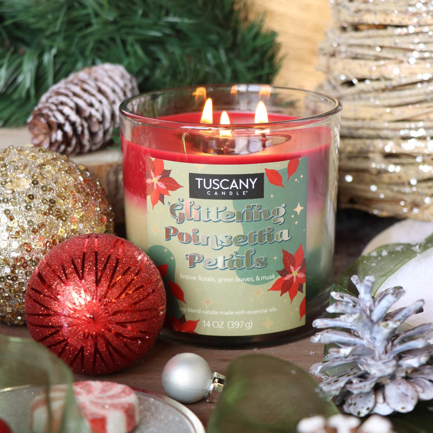 A Tuscany Candle® SEASONAL Glittering Poinsettia Petals Scented Holiday Candle (14 oz) burns with three flames, nestled among Christmas ornaments, greenery, and festive holiday décor.