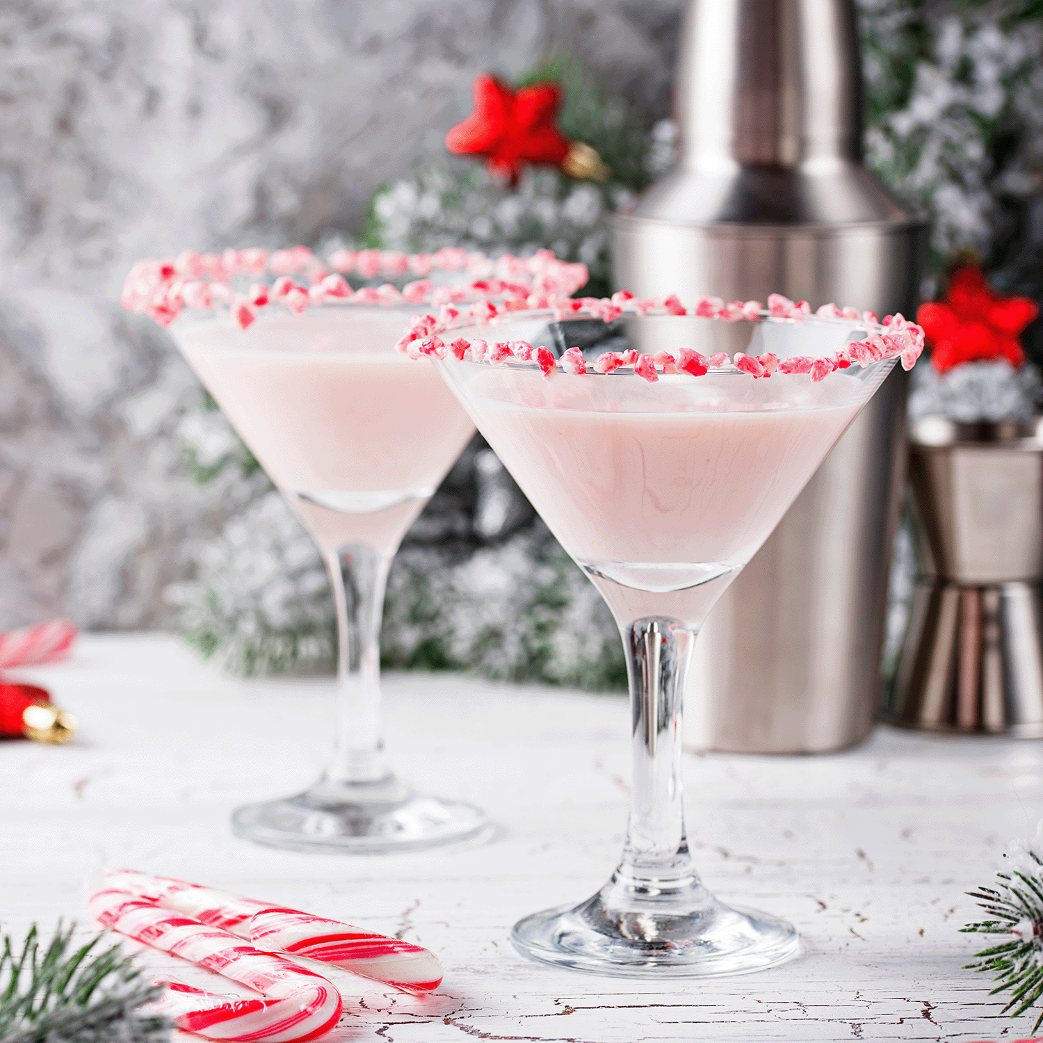 Two pink cocktails in martini glasses with crushed candy cane rims sit on a white table with holiday décor, bar tools, and the cozy aroma of Tuscany Candle® SEASONAL's Candy Cane Forest Peppermint & Balsam Holiday Wax Melt in the background.