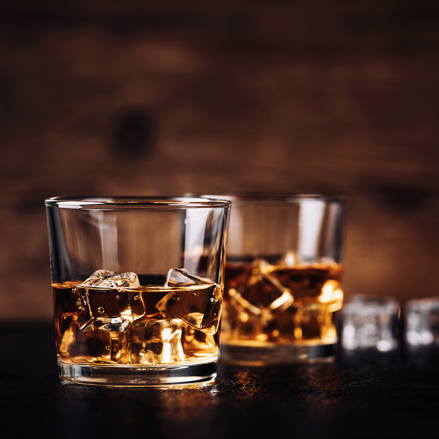 Two glasses of whiskey with ice on a dark surface, set against a blurred brown background, recall the cozy feel of Tuscany Candle® SEASONAL’s Holiday Spirits: White Rum & Cognac Scented Holiday Wax Melt (2.5 oz), filling your home with long-lasting fragrance.