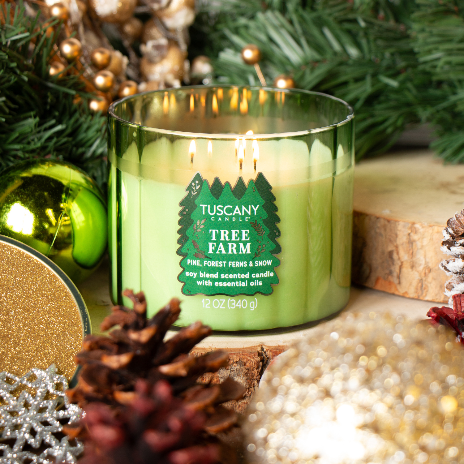 A lit Tuscany Candle® SEASONAL "Tree Farm: Forest Ferns & Snow" 12 oz candle from the Winter Icons Collection rests among pine branches, pinecones, and gold ornaments.
