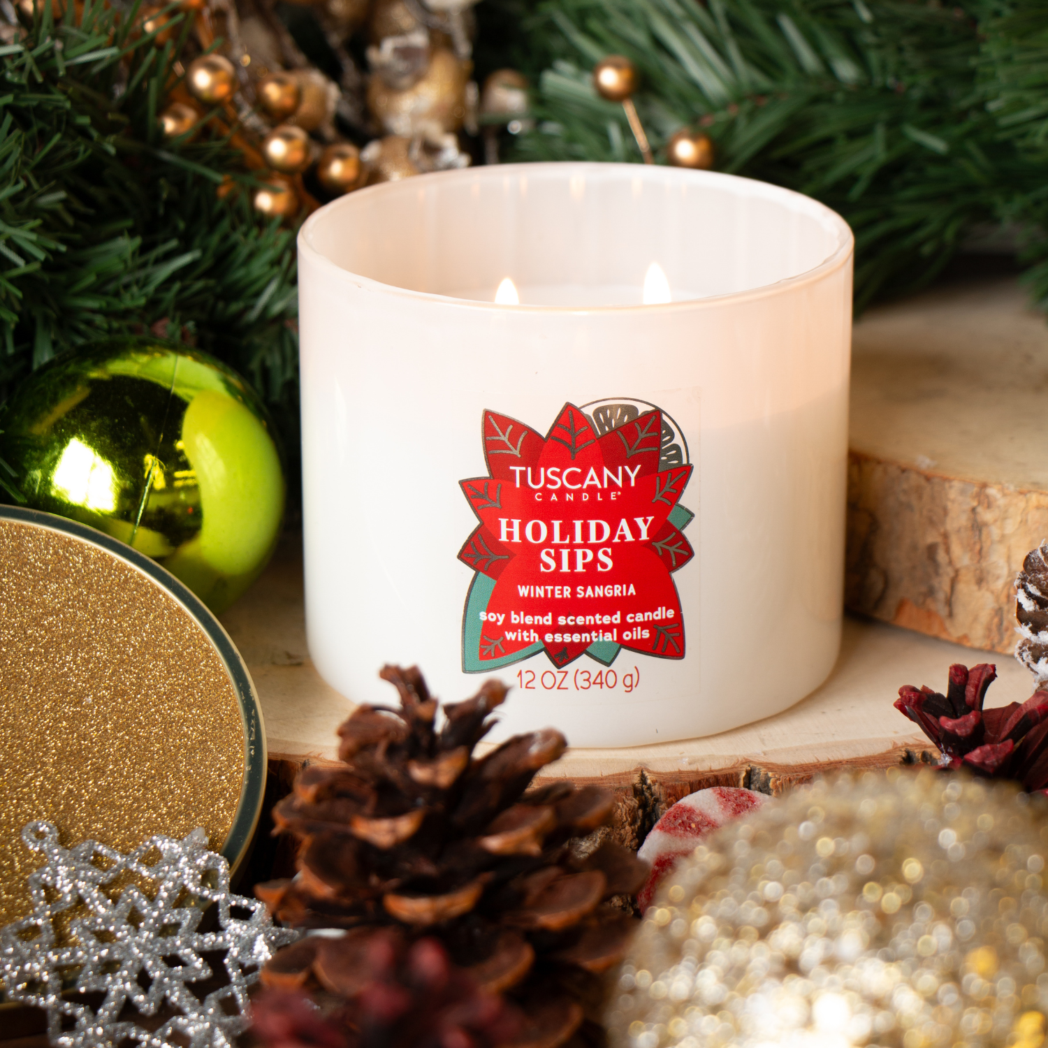 The Tuscany Candle® SEASONAL "Holiday Sips: Winter Sangria Scented Holiday Candle" (12 oz) from the Winter Icons Collection sits amid pine cones and ornaments, adding a festive touch to your holiday decor.