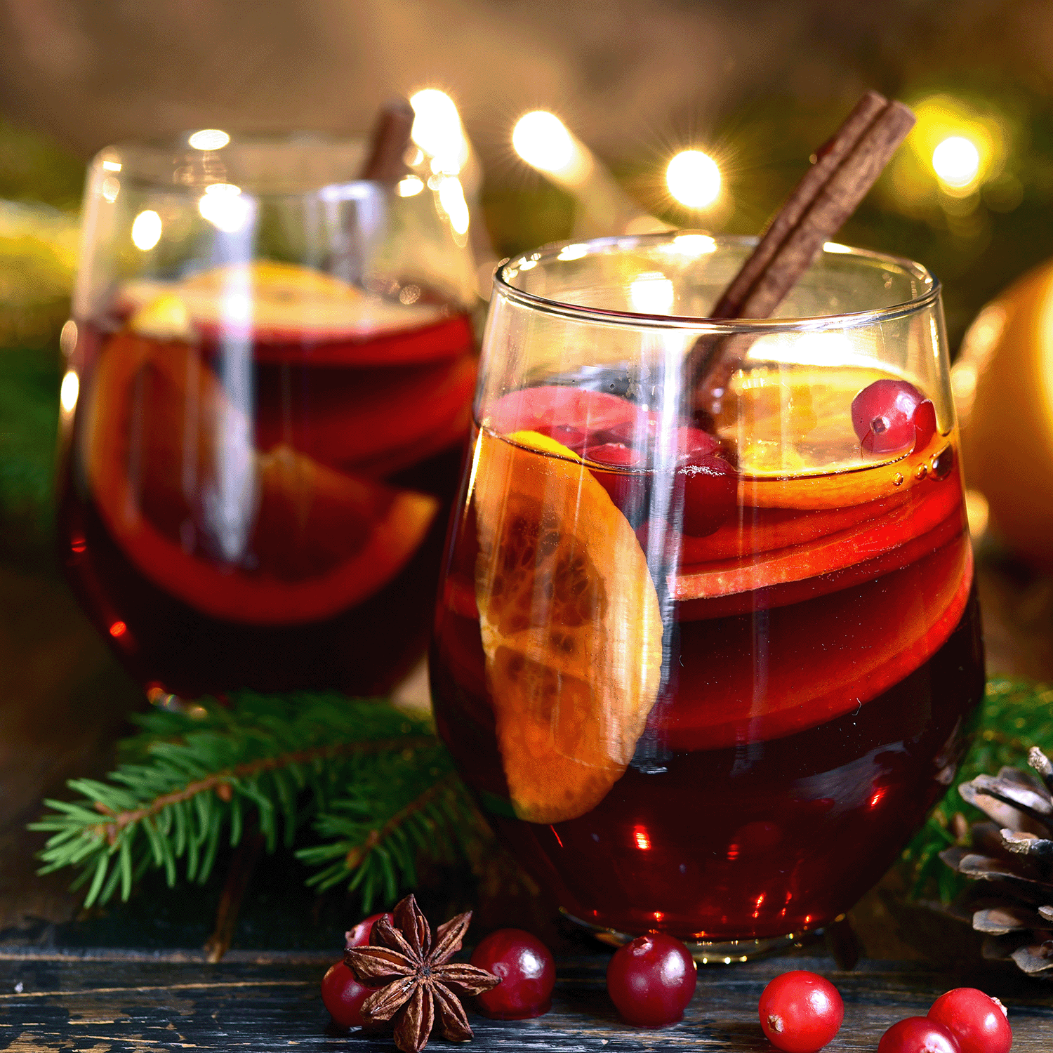 Two glasses of mulled wine rest on a wooden surface with orange, cranberries, and cinnamon, surrounded by pine and lights—the perfect scene for lighting Tuscany Candle® SEASONAL's Celebrate The Season: Winterberry & Mandarin Holiday Candle (15 oz).