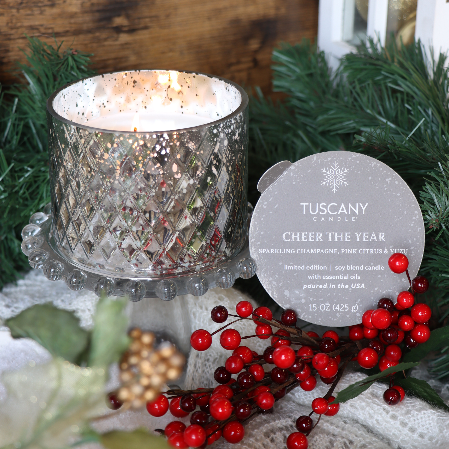 A Tuscany Candle® SEASONAL "Cheer The Year: Champagne & Yuzu" scented holiday candle (15 oz, Winter Glow Collection) glows in a decorative glass holder beside red berries and greenery.