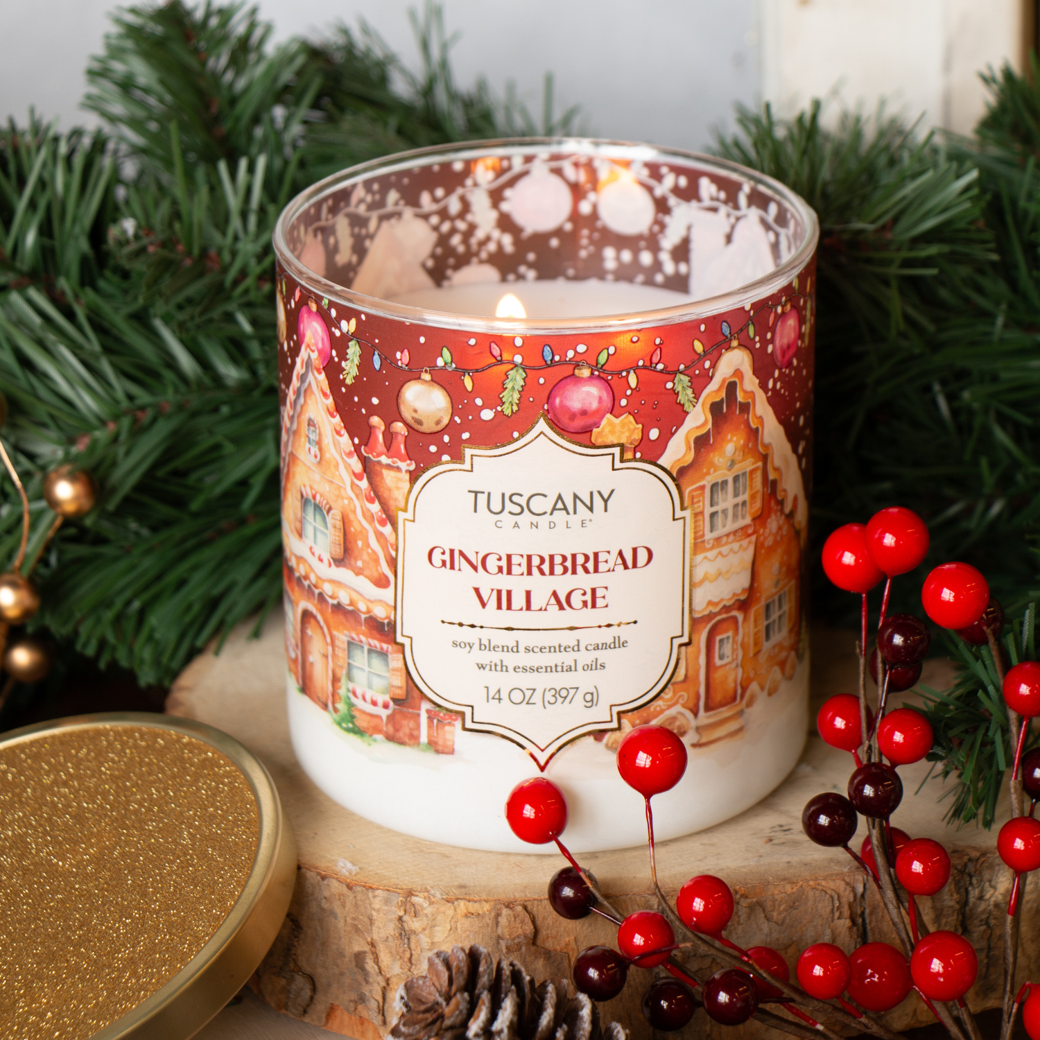 A Tuscany Candle® SEASONAL Gingerbread Village: Classic Gingerbread & Sugar Holiday Candle (14 oz) from the Traditions Collection sits near pine branches and red berries, its gold lid beside it on a wooden surface.