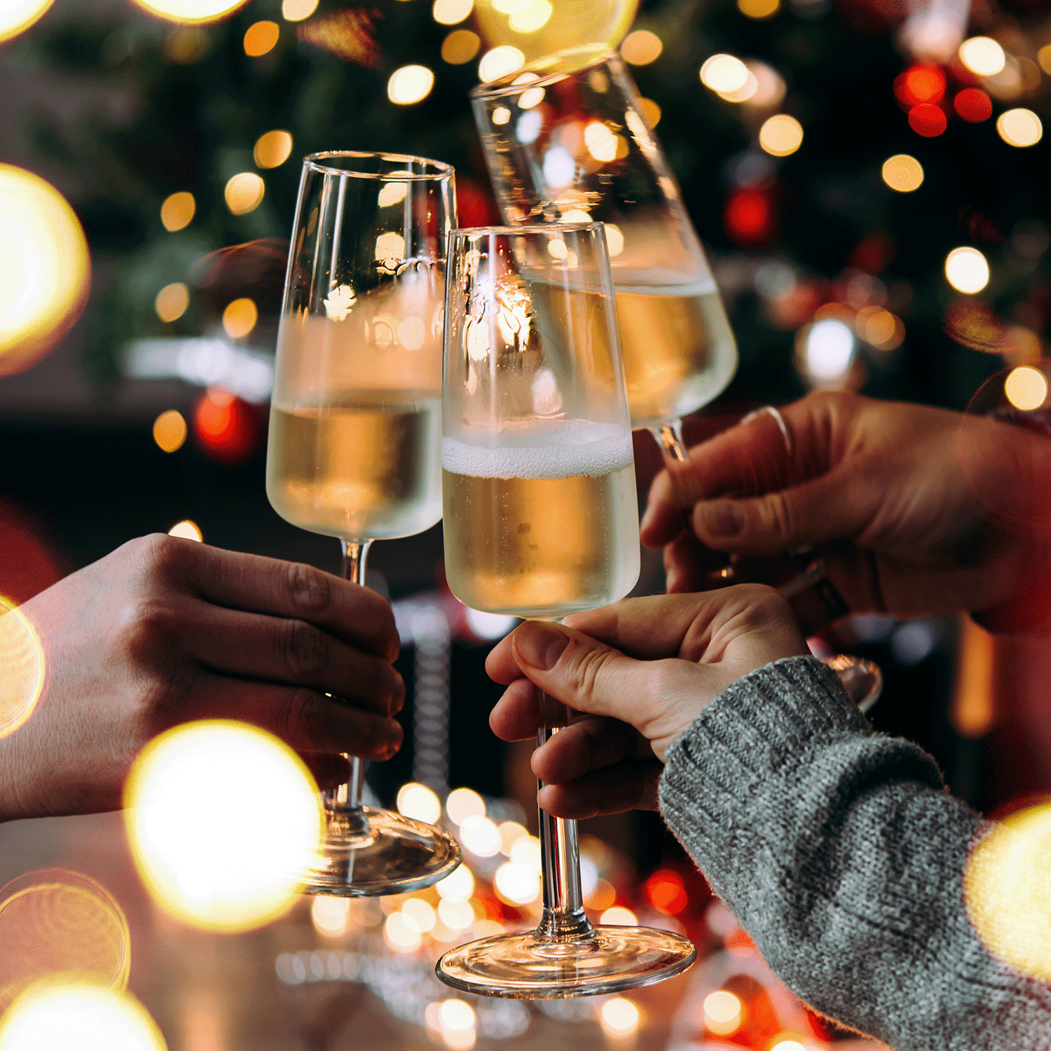 Four people toast with champagne glasses amid festive lights and decor, illuminated by the warm glow of Tuscany Candle® SEASONAL's Pink Citrus Champagne: Grapefruit & Pear Scented Holiday Candle (12 oz) from the Bakery Tins Collection.
