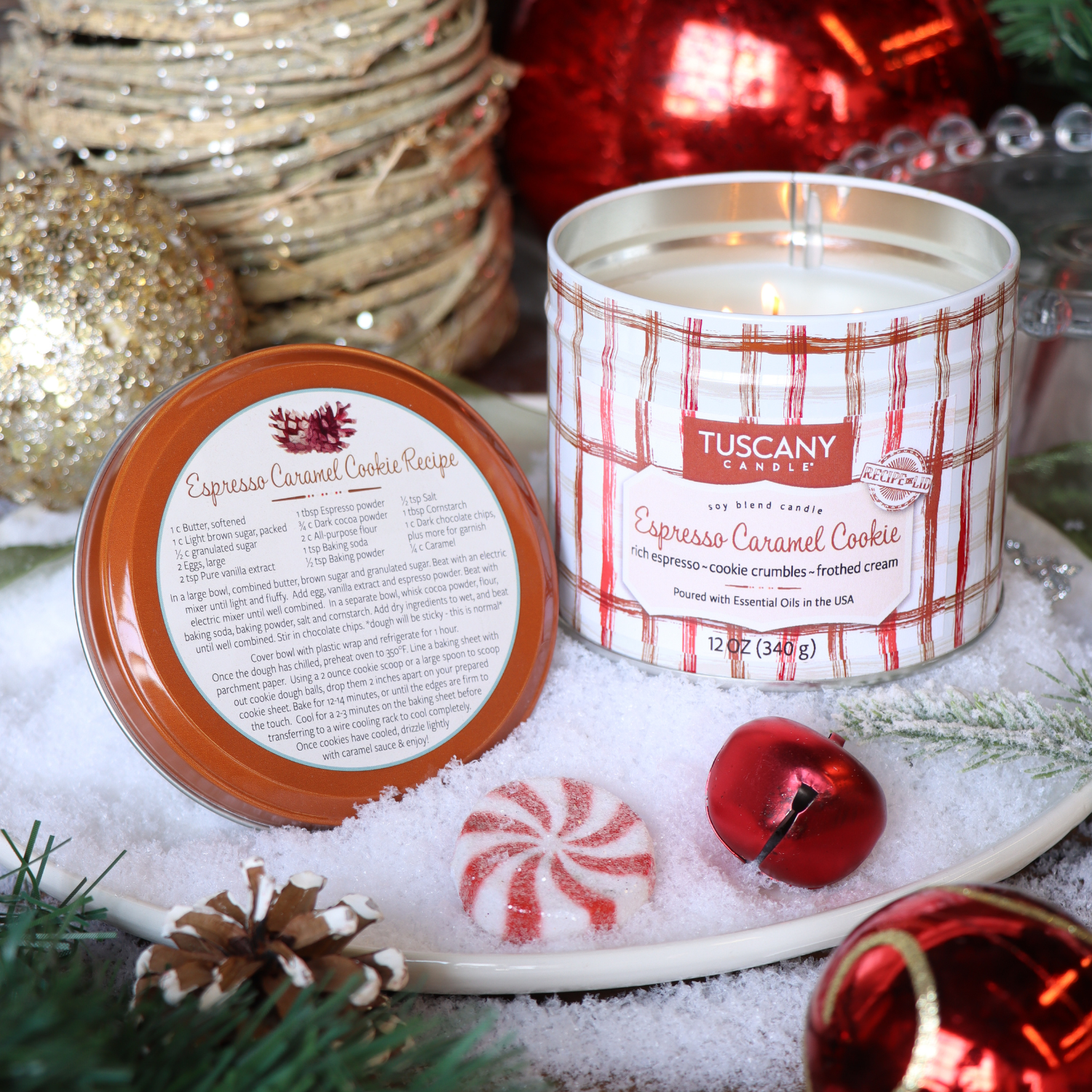 A Tuscany Candle® SEASONAL “Espresso Caramel Cookie: Sea Salt & Vanilla Bean Scented Holiday Candle” (12 oz, Bakery Tins Collection) sits open on artificial snow amid holiday decor and greenery, with its lid displaying a cookie recipe.