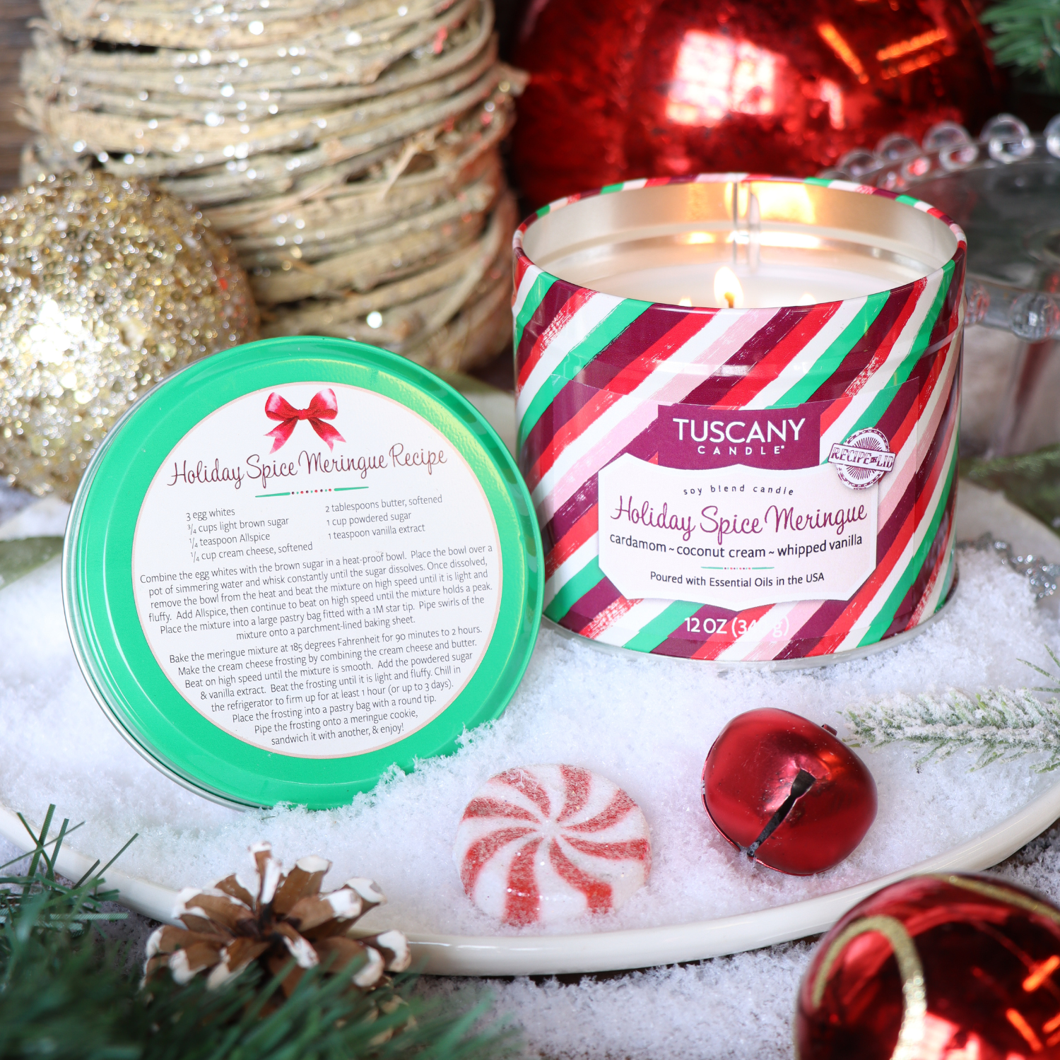 A Tuscany Candle® SEASONAL "Holiday Spice Meringue: Cardamom & Vanilla Scented Holiday Candle" (12 oz, Bakery Tins Collection) sits open on a snowy surface with its lid showing a holiday spice meringue recipe, surrounded by festive décor.