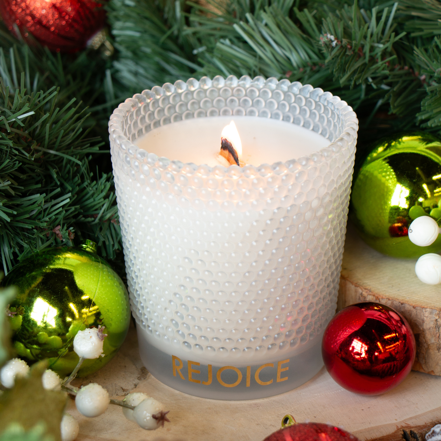 A Tuscany Candle® SEASONAL "Rejoice" 13 oz wooden wick candle, with peppermint & vanilla scent, sits in a textured glass holder amid Christmas ornaments, greenery, and berries.