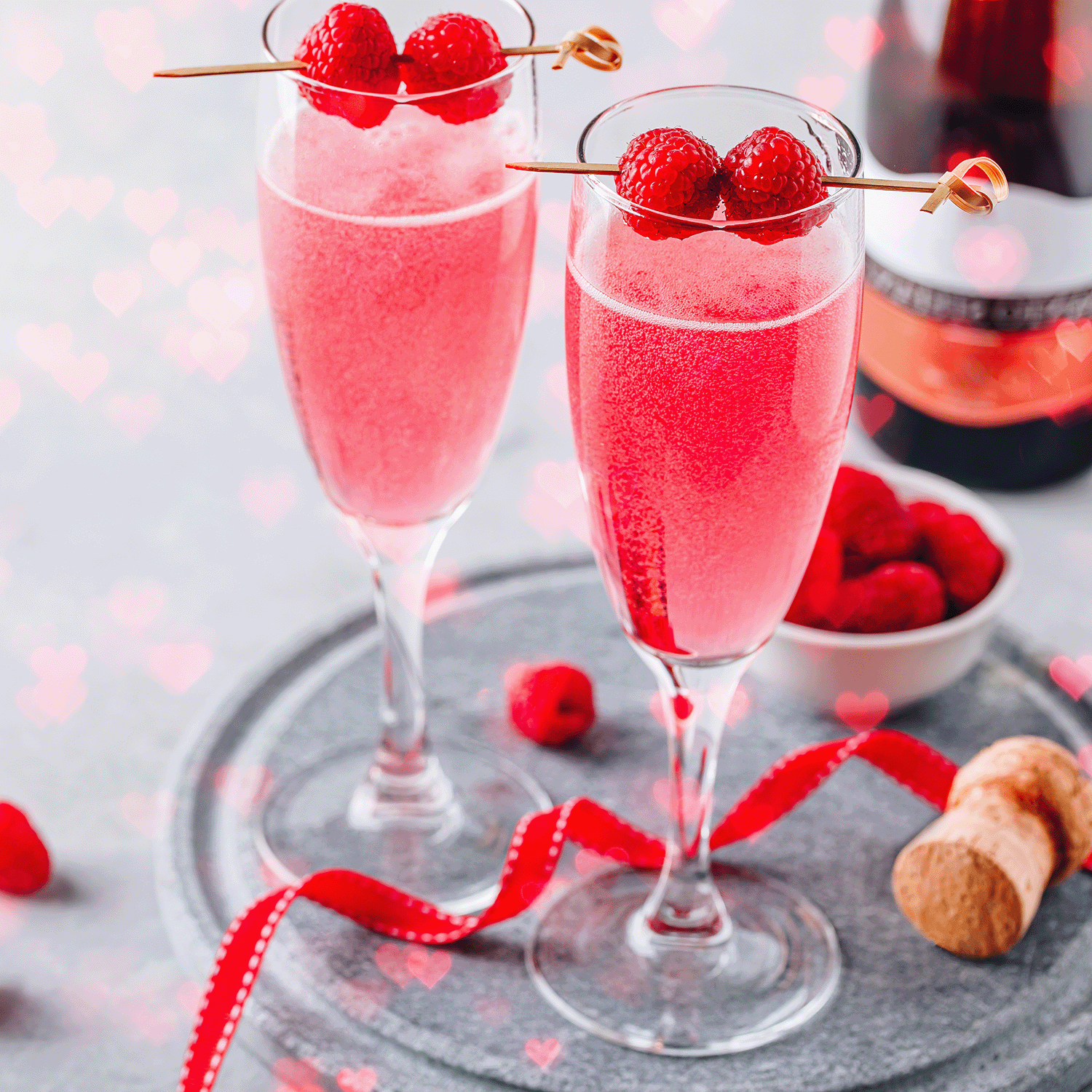 Two champagne flutes filled with pink sparkling drink, topped with raspberries on skewers, sit on a tray beside Tuscany Candle® SEASONAL's Rosé & Roses: Pink Rose Scented Valentine's Wax Melt (2.5 oz), with ribbon and loose raspberries.