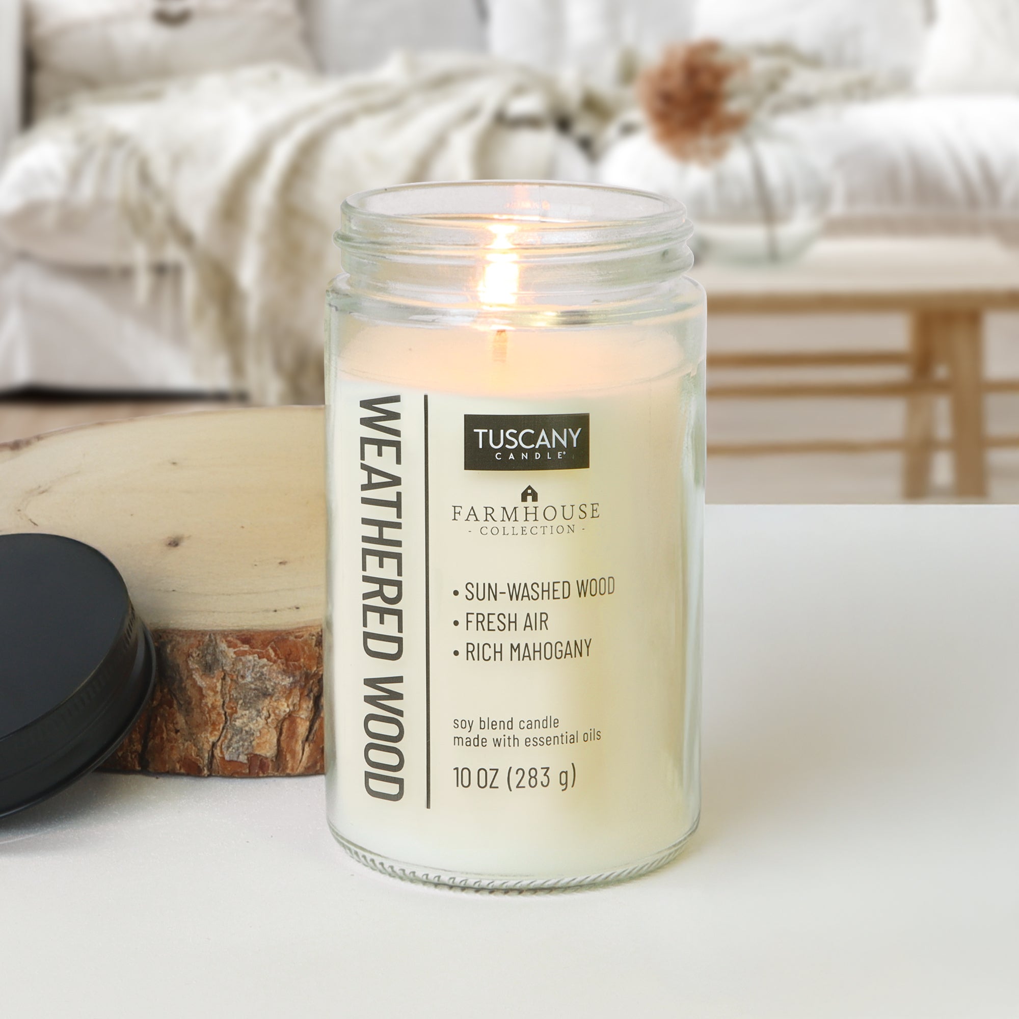 A glowing Weathered Wood Scented Jar Candle (10 oz) from the Tuscany Candle® EVD Farmhouse Collection shines in a clear glass jar on wood, with a cozy couch and blanket softly blurred in the background.