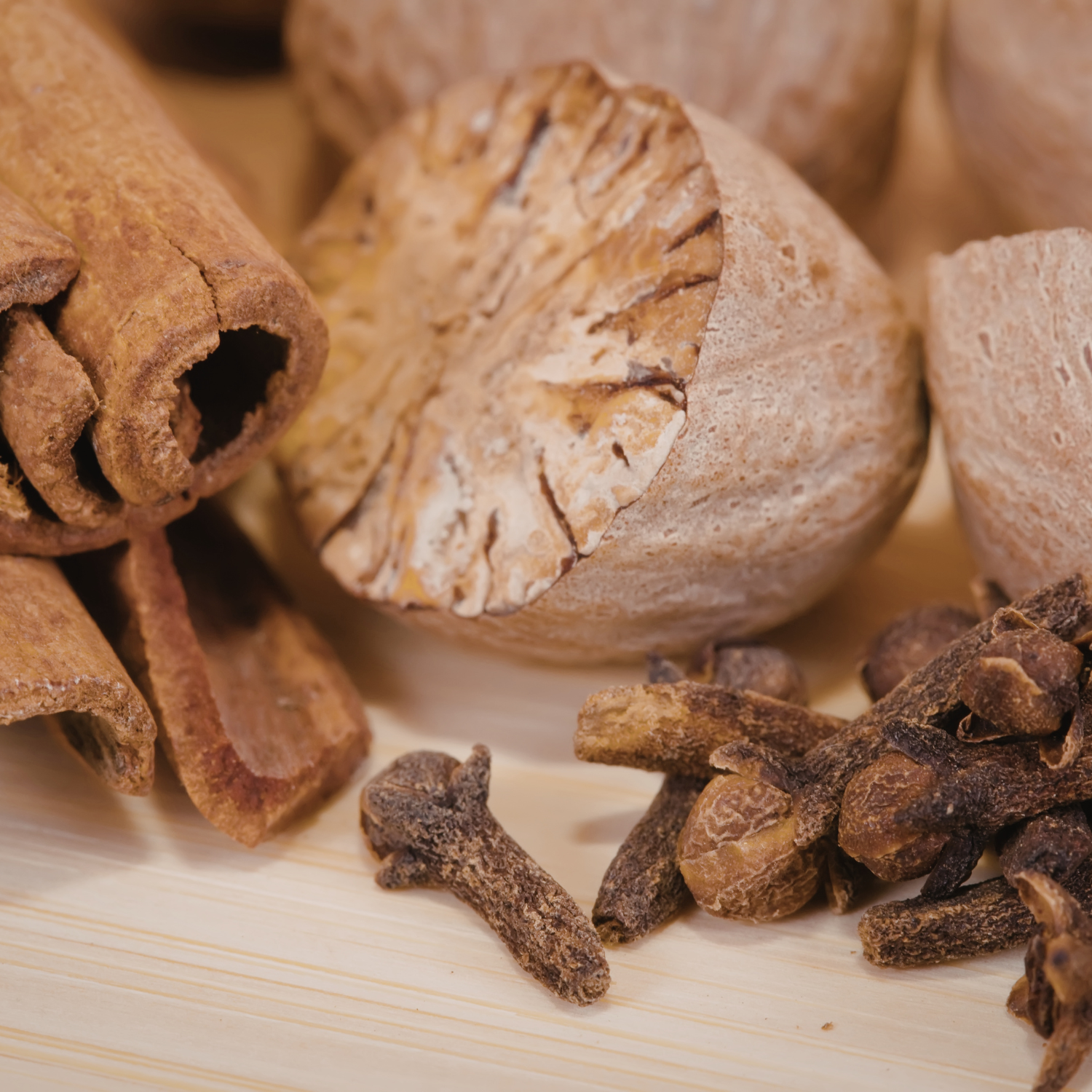 Close-up of whole nutmeg, cinnamon sticks, and cloves on a light wooden surface beside a Tuscany Candle® EVD Comforts of Home Scented Jar Candle (10 oz) from the Farmhouse Collection.