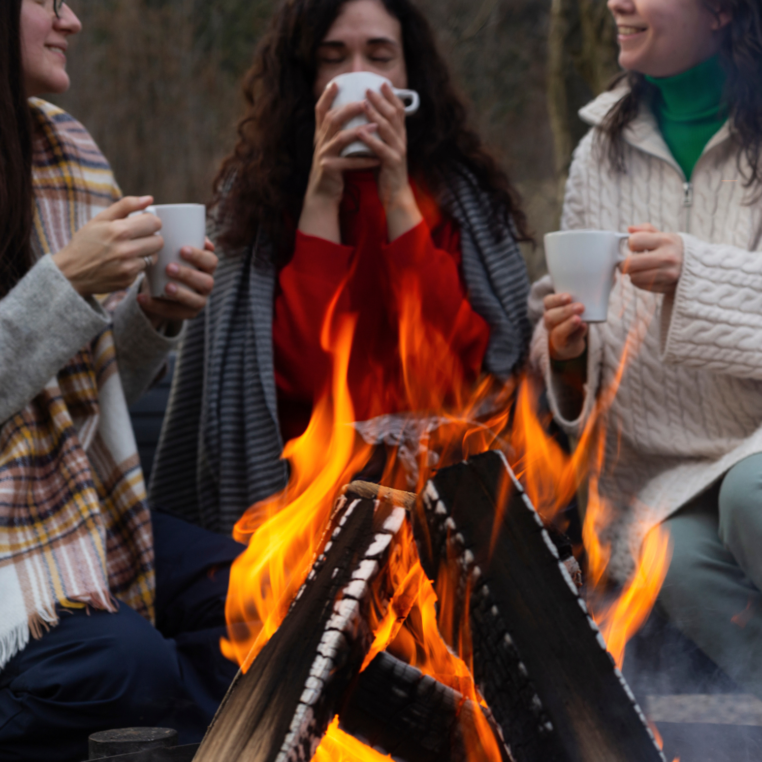 Three friends gather by a glowing campfire, mugs in hand and warm clothes on, evoking cozy farmhouse vibes—just like the inviting ambiance of the Tuscany Candle® EVD Fireside Scented Jar Candle (10 oz) from the Farmhouse Collection.