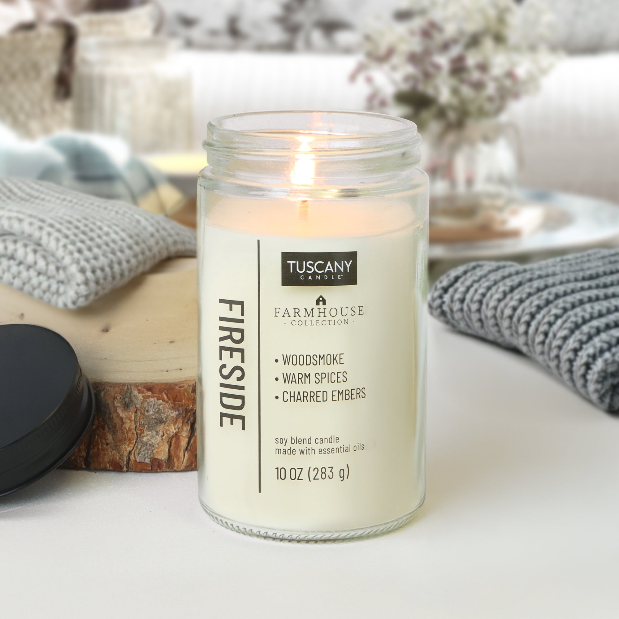A lit Tuscany Candle® EVD Fireside Scented Jar Candle (10 oz) from the Farmhouse Collection sits on a table with folded blankets and a fireplace, filling the room with cozy warmth and enhancing farmhouse decor.