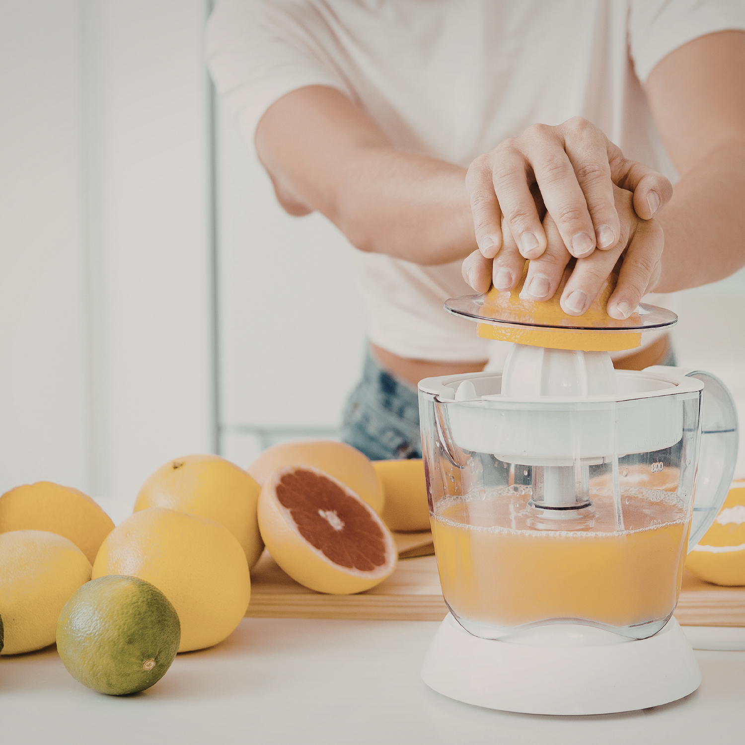 A person uses a citrus juicer to extract juice from an orange, while a Tuscany Candle® EVD Crisp Apple Scented Jar Candle (10 oz) from the Farmhouse Collection glows nearby amid lemons, limes, and a halved grapefruit on the white countertop.