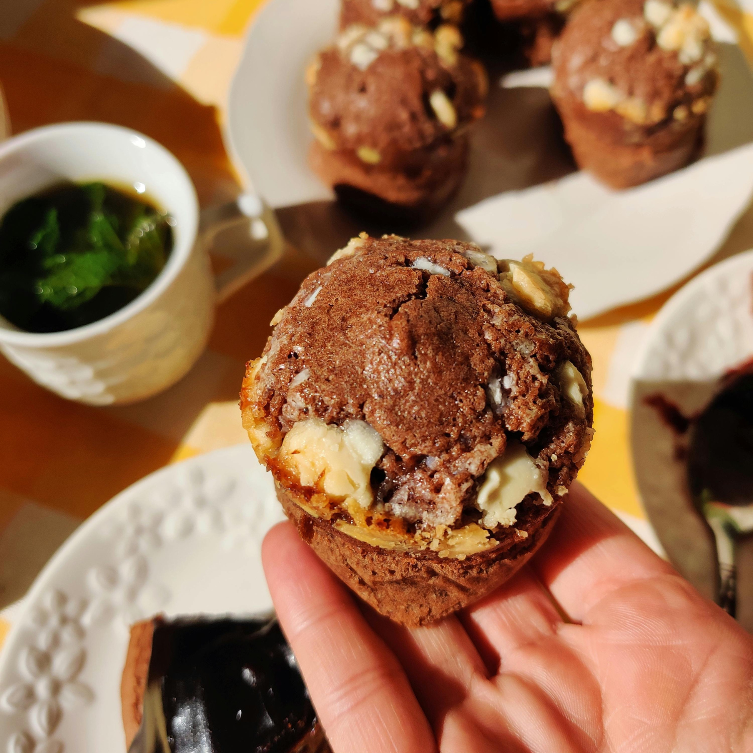 A hand holds a chocolate muffin with nuts; more muffins, a cup of tea, a Tuscany Candle® EVD Homemade Muffins Scented Jar Candle (10 oz) – Farmhouse Collection, and a chocolate dessert are on the table in the background.