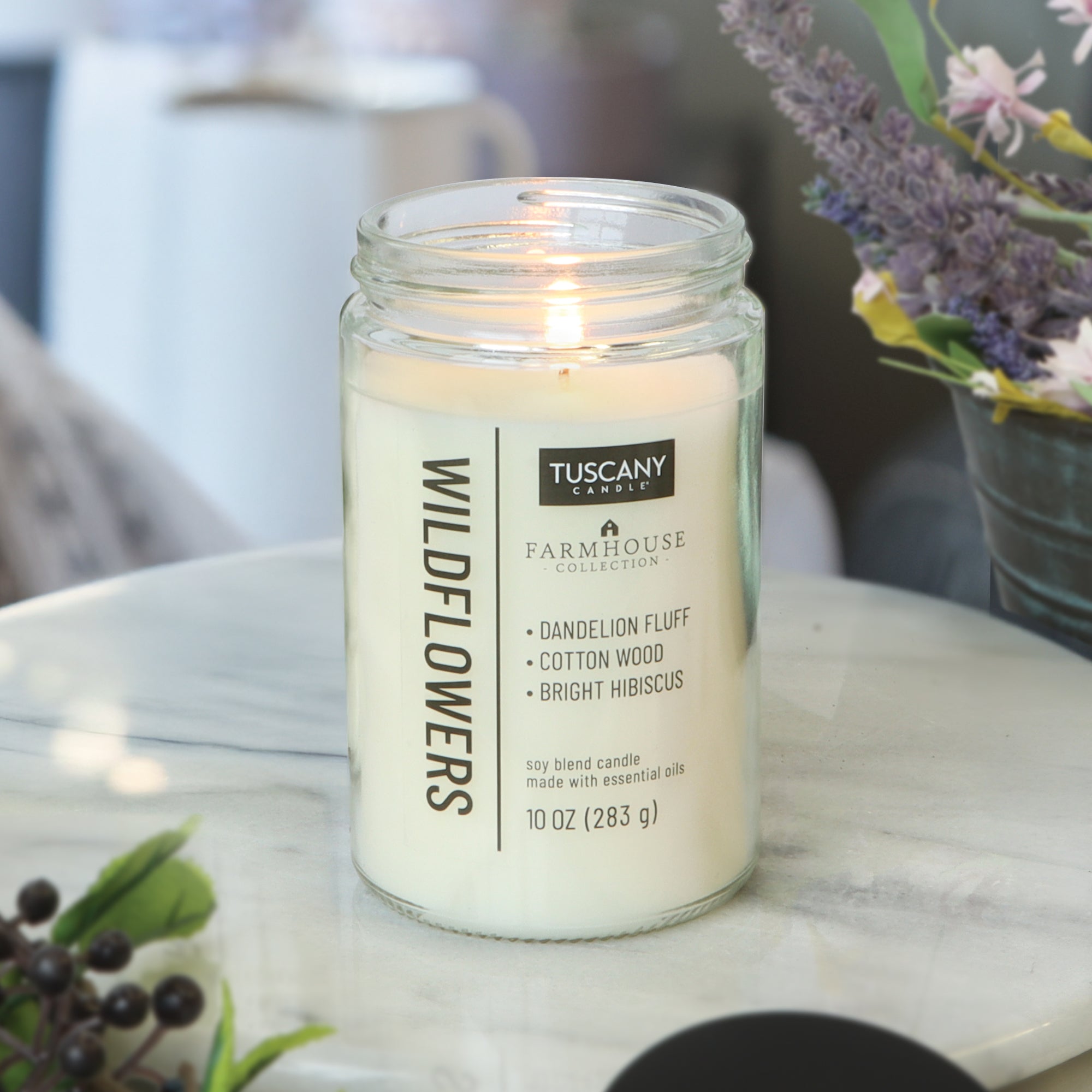A lit Wildflowers Scented Jar Candle (10 oz) from the Tuscany Candle® EVD Farmhouse Collection sits on marble, filling the room with notes of dandelion fluff, cottonwood, and bright hibiscus—ideal for a farmhouse-inspired space.