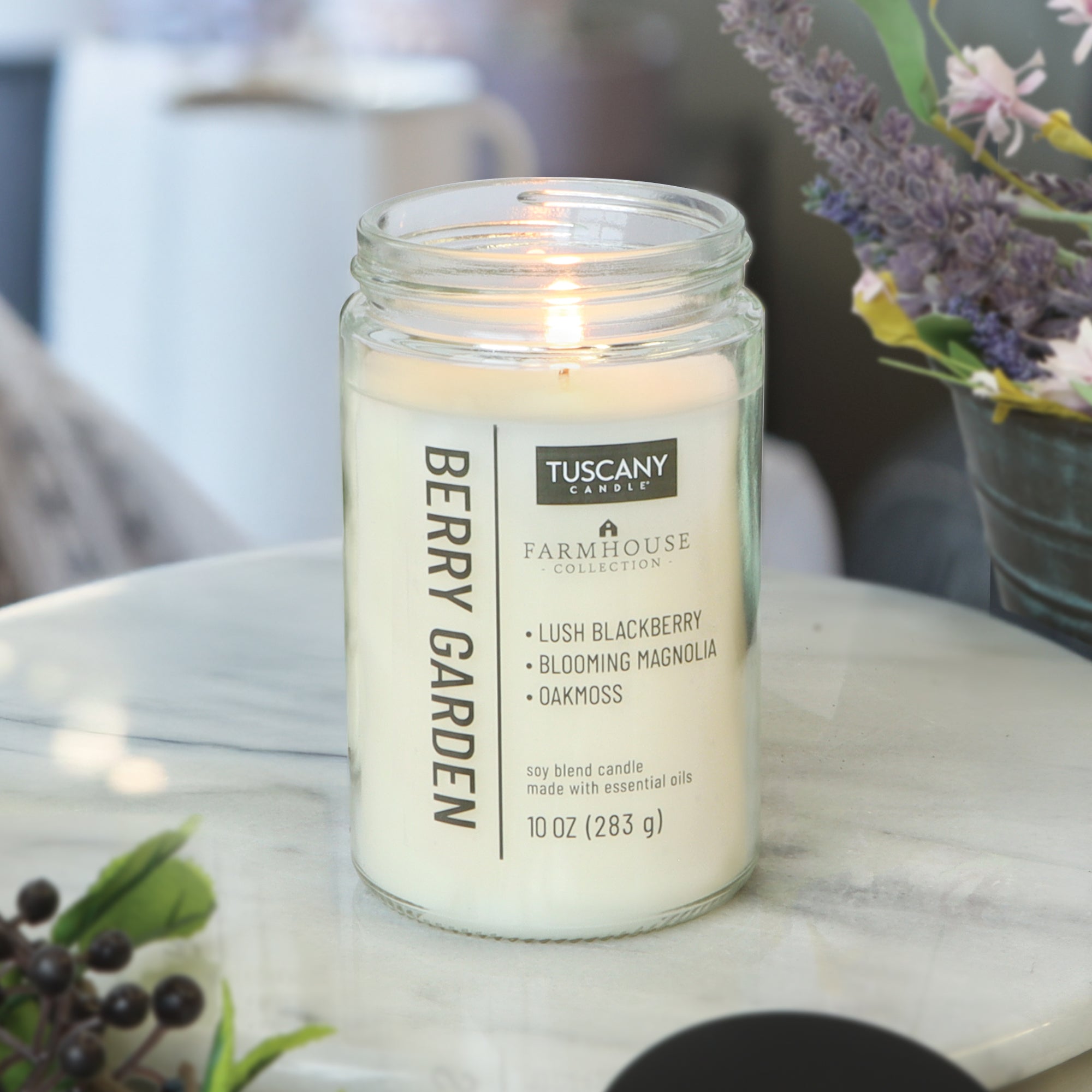 A Berry Garden Scented Jar Candle (10 oz) from the Tuscany Candle® EVD Farmhouse Collection glows on a marble table among flowers and berries, featuring lush blackberry, blooming magnolia, and oakmoss scents.