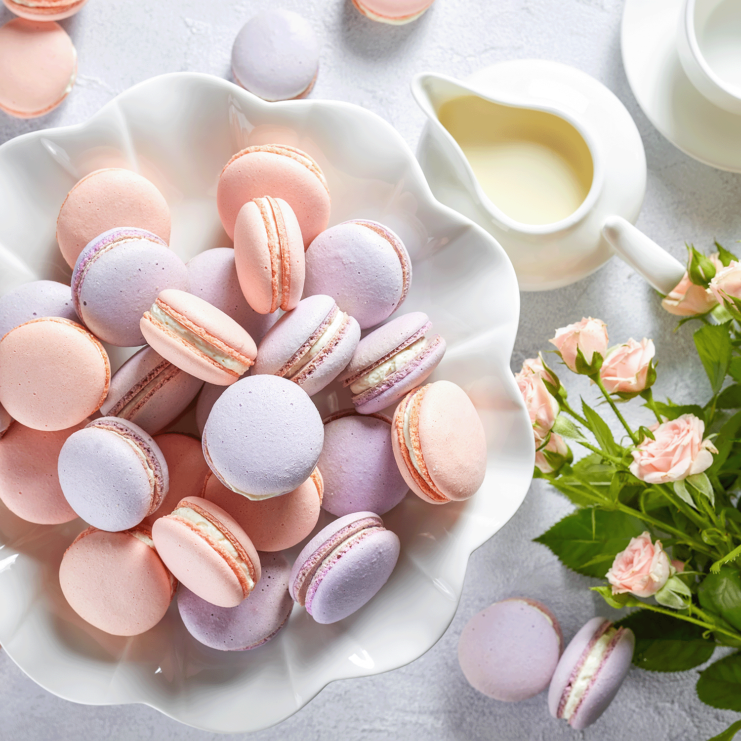 A white bowl of pink and purple macarons sits beside the Tuscany Candle® SEASONAL Lavender Macaron: Vanilla Bean & Sugar Scented Spring Candle (14 oz), a small cream pitcher, pink roses, and scattered macarons on the table.