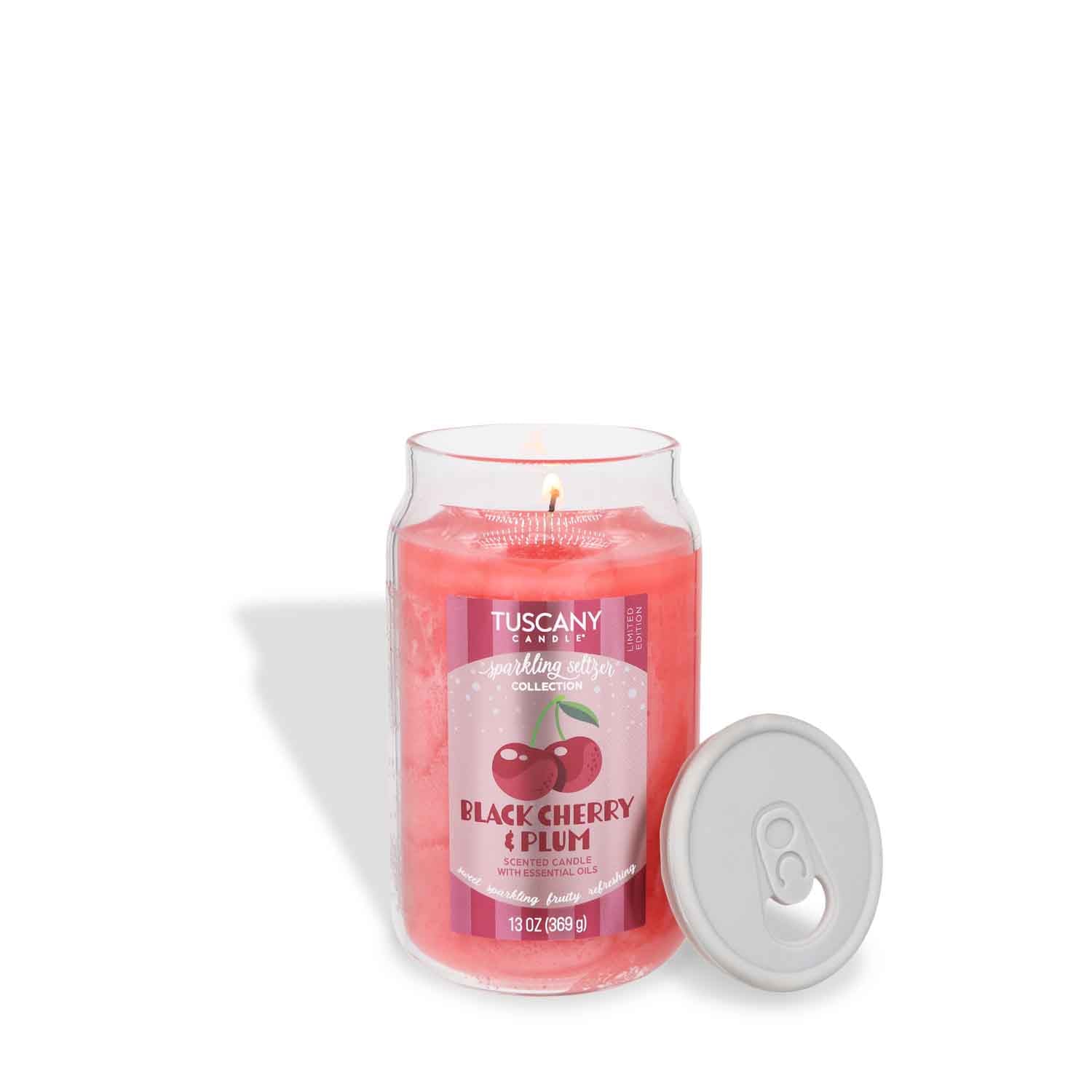 A lit 13 oz Tuscany Candle® SEASONAL “Black Cherry & Plum: Sparkling Seltzer Scented Summer Candle” sits beside its gray lid on a white background.