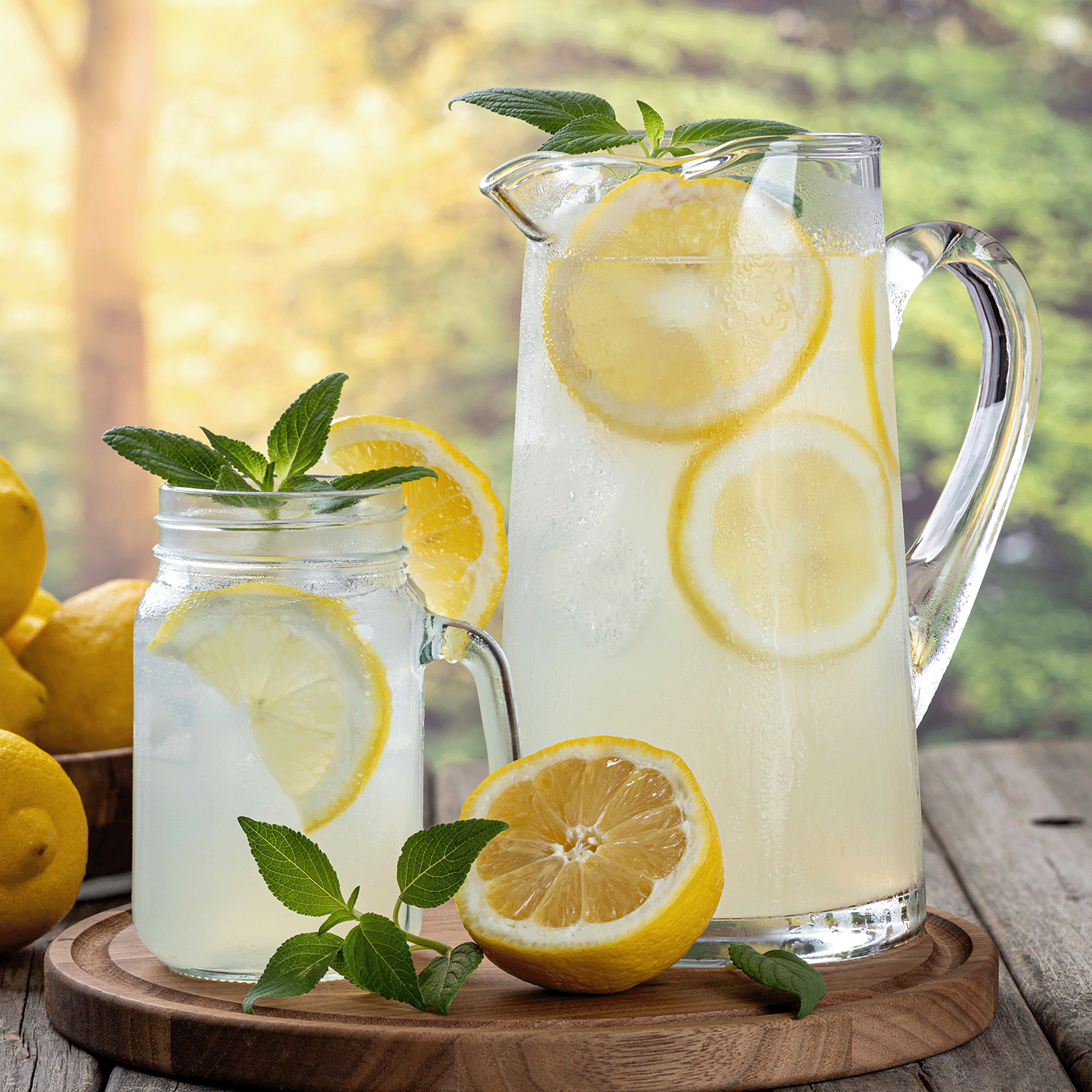 A glass pitcher and mason jar of lemonade with lemon slices and mint on a wooden tray, surrounded by lemons and the Tuscany Candle® SEASONAL Limoncello Fruit Slices: Meyer Lemon Scented Summer Candle (12 oz) for a perfect summer scene.