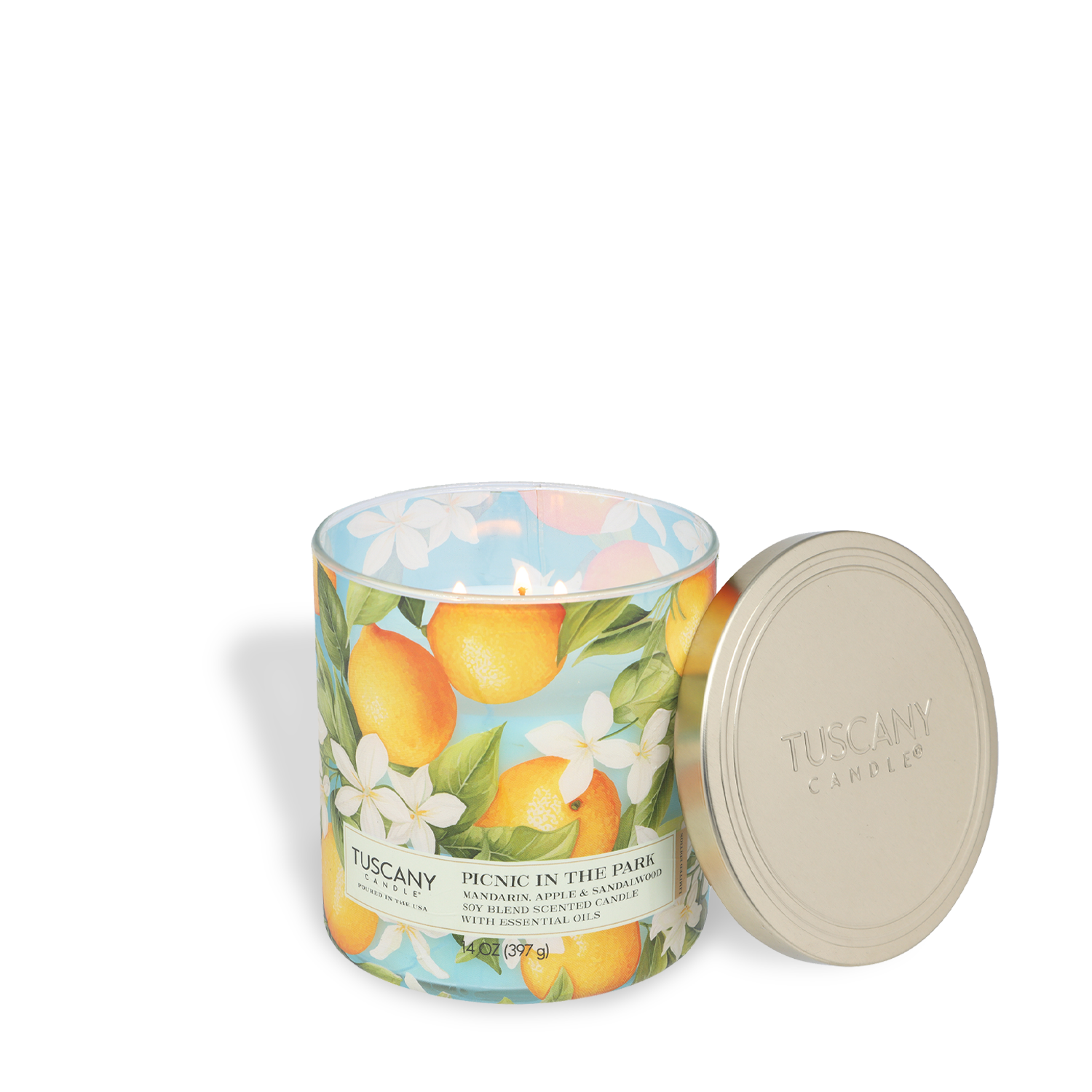 A lit Tuscany Candle® SEASONAL "Picnic In The Park: Mandarin & Mango" 14 oz soy blend candle from the Dreamy Destinations Collection sits in a glass jar with floral-citrus design; its metal lid rests to the side.