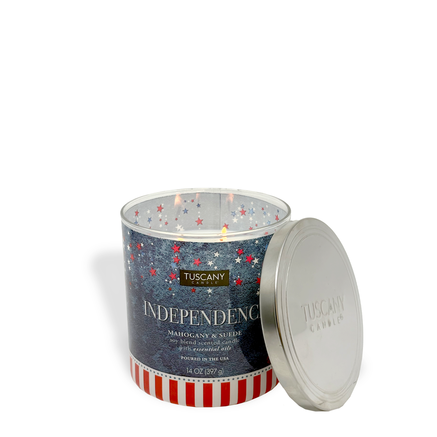 The lit Tuscany Candle® SEASONAL "Independence: Mahogany & Suede Scented Summer Candle" (14 oz) sits in a glass jar with stars and stripes, its lid resting beside it—a perfect patriotic home fragrance for summer.