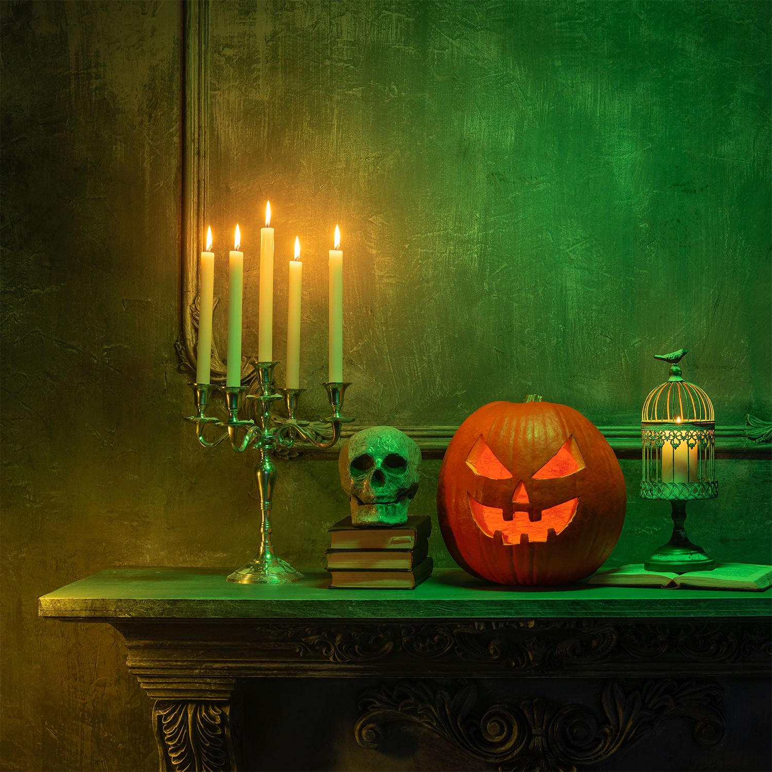 A Wicked Pumpkins: Caramel & Pumpkin Scented Halloween Wax Melt by Tuscany Candle® SEASONAL, a skull resting on books, a candelabra, and a birdcage are displayed on a table against a green-lit wall in a Halloween-themed setting.