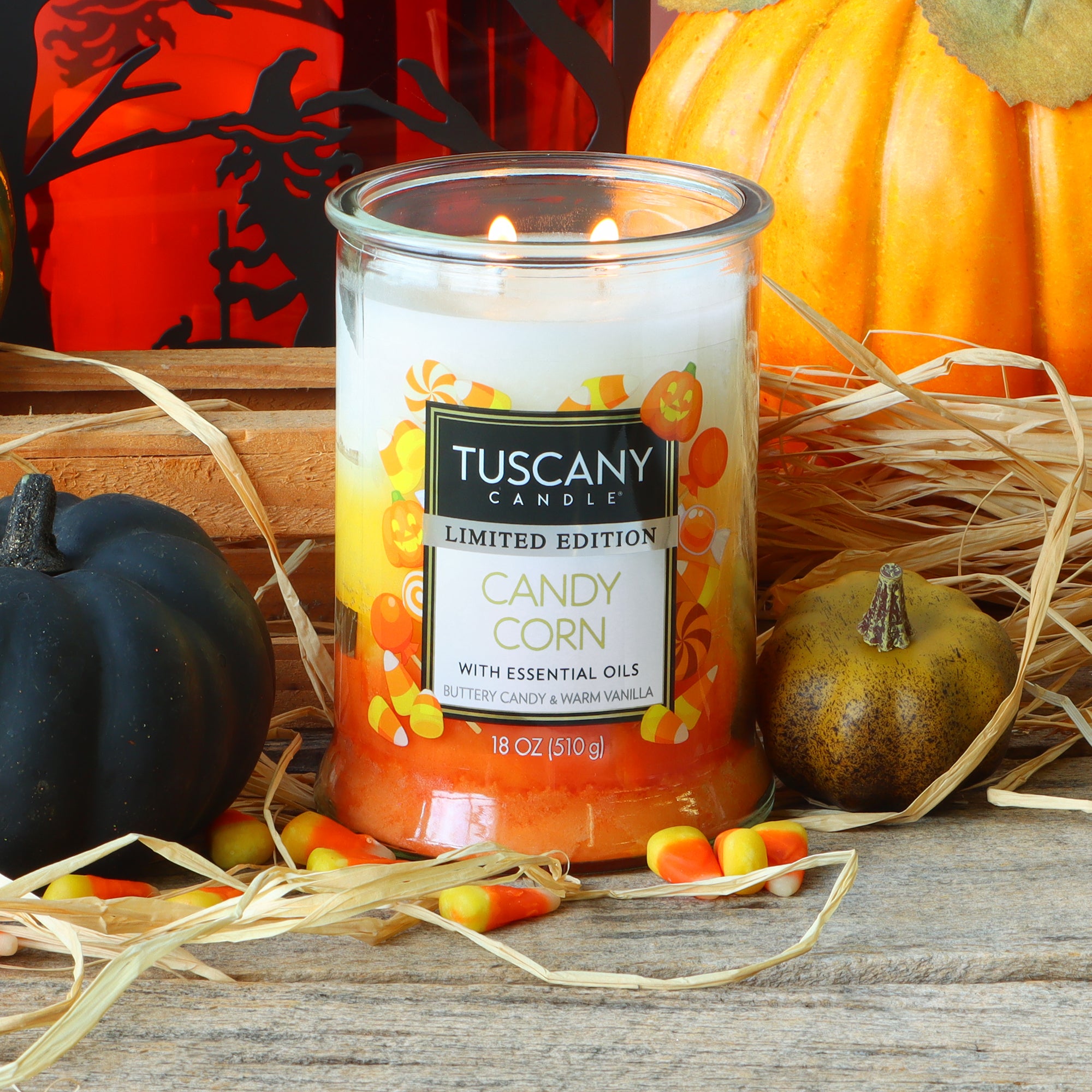 The Tuscany Candle® SEASONAL “Candy Corn: Buttery Candy & Vanilla Scented Halloween Candle” (18 oz) is lit and surrounded by candy corn pieces, hay, pumpkins, and gourds on a wooden surface.