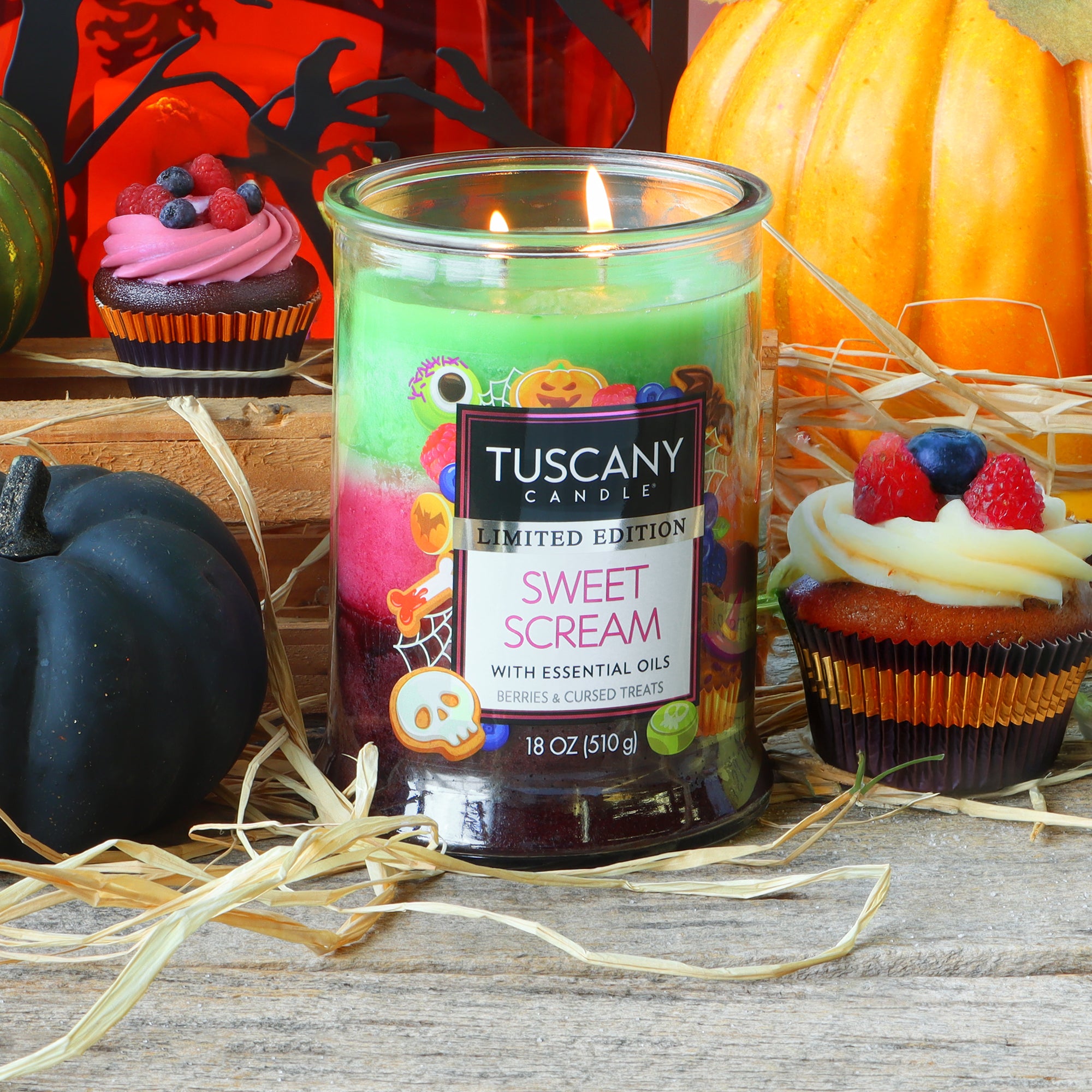 An 18 oz Tuscany Candle® SEASONAL “Sweet Scream: Berries & Cursed Treats” scented Halloween candle burns on a wooden table surrounded by cupcakes, a straw, pumpkins, and festive Halloween decorations.