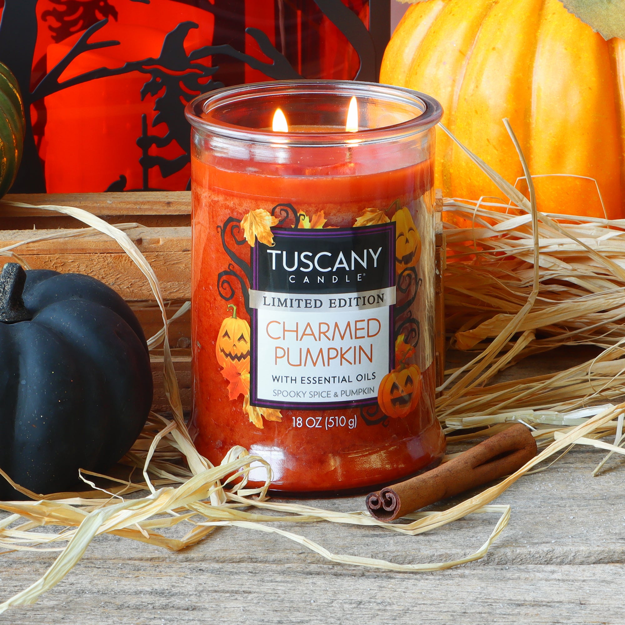 A glowing Tuscany Candle® SEASONAL “Charmed Pumpkin: Spooky Spice & Pumpkin Scented Halloween Candle” (18 oz) rests on a wooden surface, surrounded by hay, cinnamon sticks, and pumpkins.