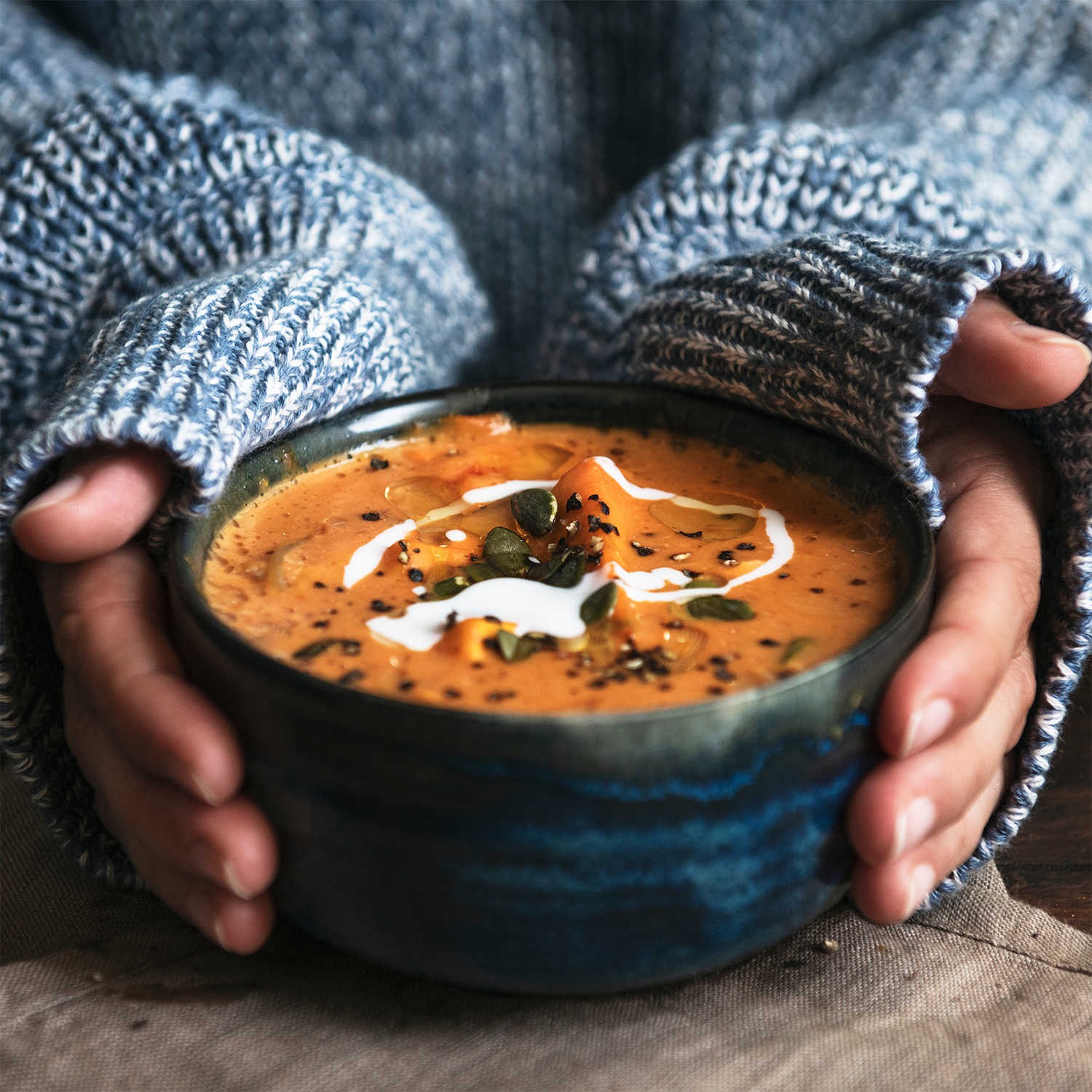Hands in a blue sweater hold a Tuscany Candle® SEASONAL Charmed Pumpkin candle, its blue ceramic vessel filled with 18 oz of creamy orange wax, topped with festive pumpkin and spooky spice scents.