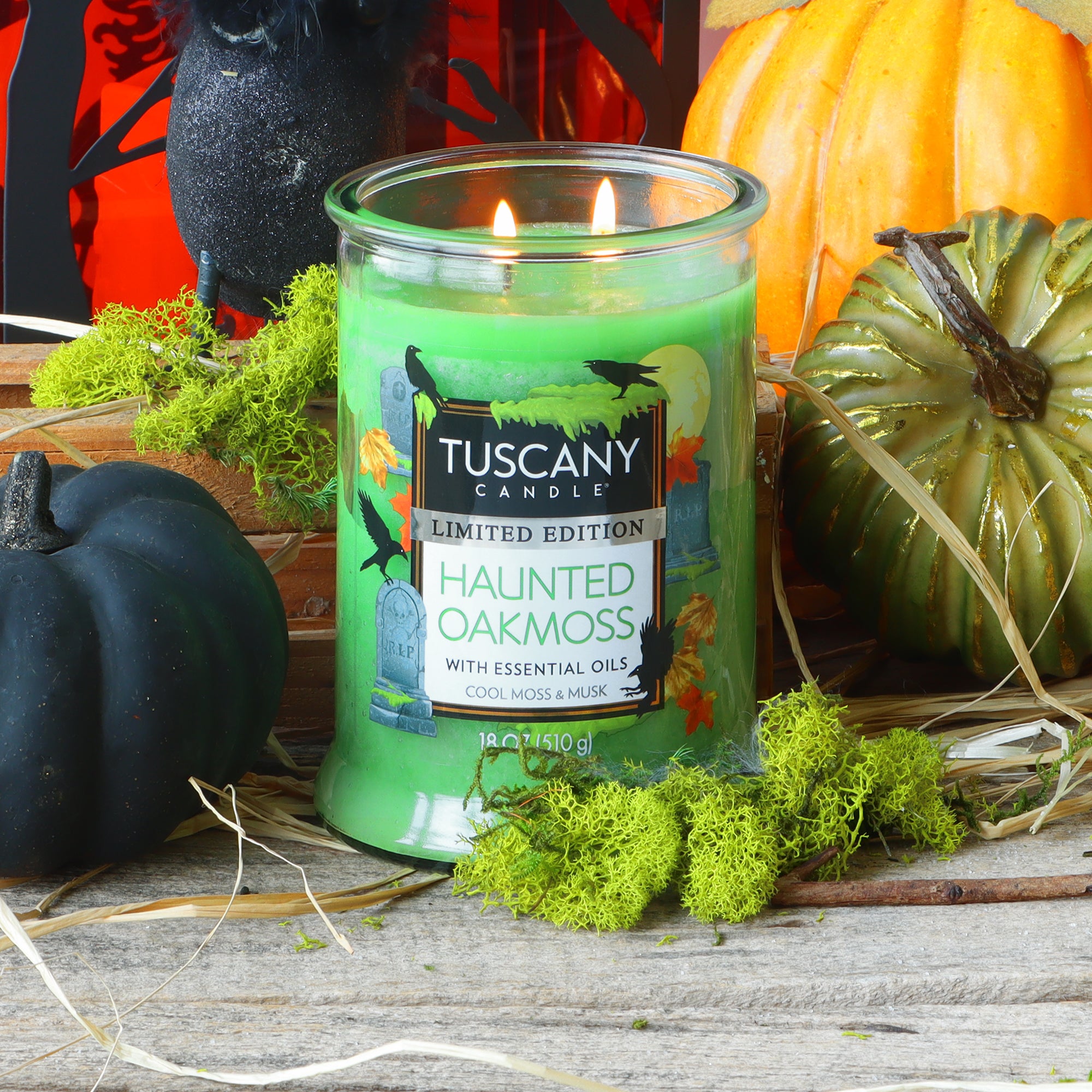 An 18 oz Tuscany Candle® SEASONAL “Haunted Oakmoss: Cool Moss & Musk Scented Halloween Candle” glows on a wooden surface, surrounded by moss, black and green pumpkins, straw, and festive Halloween decorations.