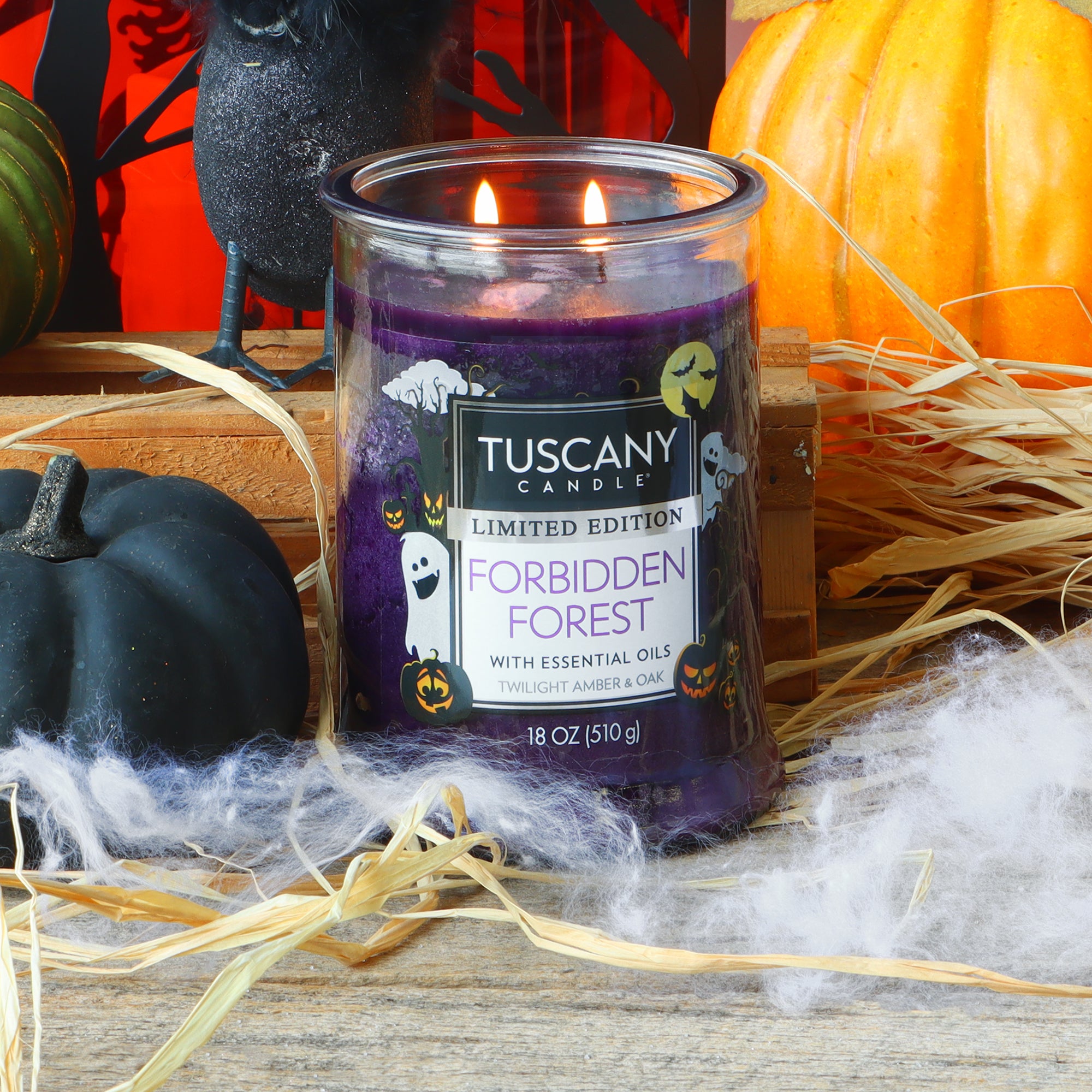 A purple Tuscany Candle® SEASONAL candle labeled “Forbidden Forest: Twilight Amber & Oak Scented Halloween Candle (18 oz)” glows with two flames, surrounded by straw, white webbing, a pumpkin, and black decorative accents in a festive Halloween display.