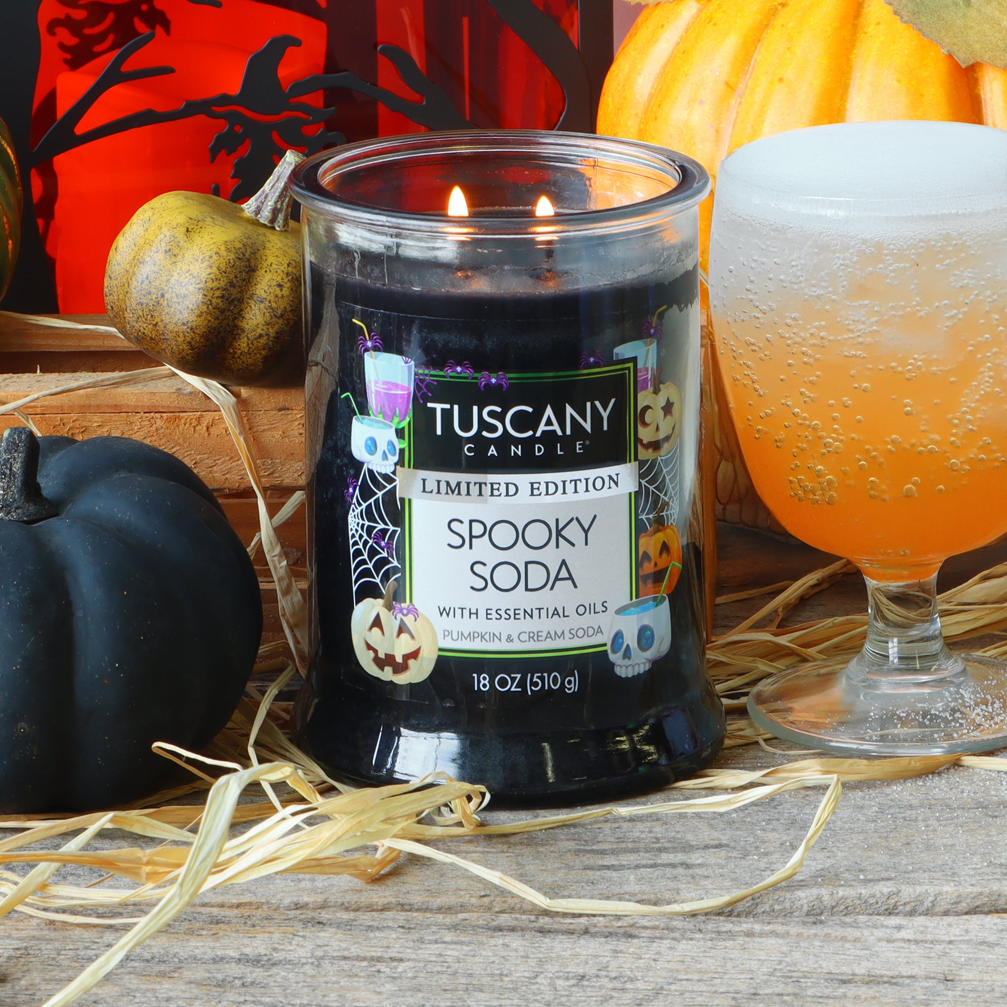 An 18 oz Tuscany Candle® SEASONAL “Spooky Soda: Pumpkin & Cream Soda Scented Halloween Candle” sits on straw next to a black pumpkin, an orange beverage in a frosted glass, a decorative pumpkin, and festive Halloween accents.
