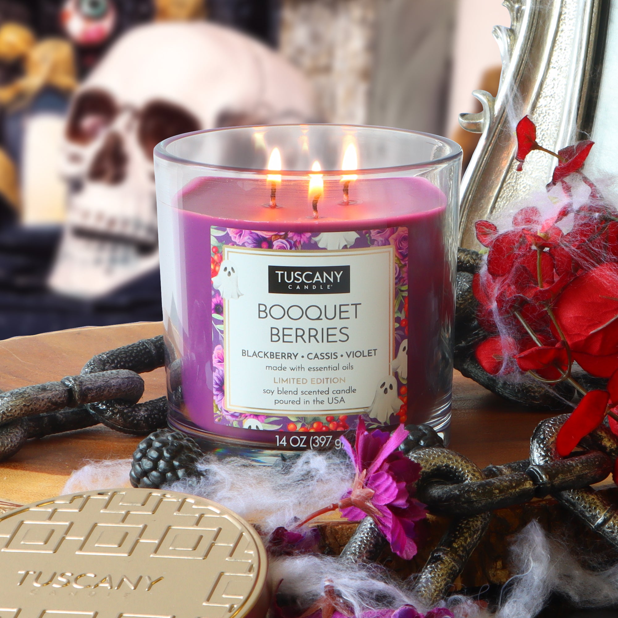 A lit Tuscany Candle® SEASONAL “Booquet Berries: Blackberry & Cassis Scented Halloween Candle” (14 oz) sits on a table decorated with Halloween items, including a skull, artificial spiderwebs, and red flowers.