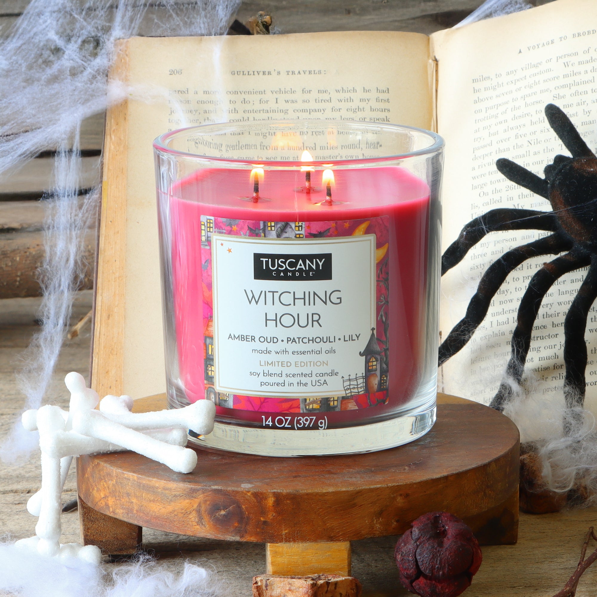 A red, three-wick Witching Hour: Patchouli & Oud Scented Halloween Candle (14 oz) by Tuscany Candle® SEASONAL sits on a wooden stand, surrounded by faux cobwebs, bones, a spider, and open books in the background.