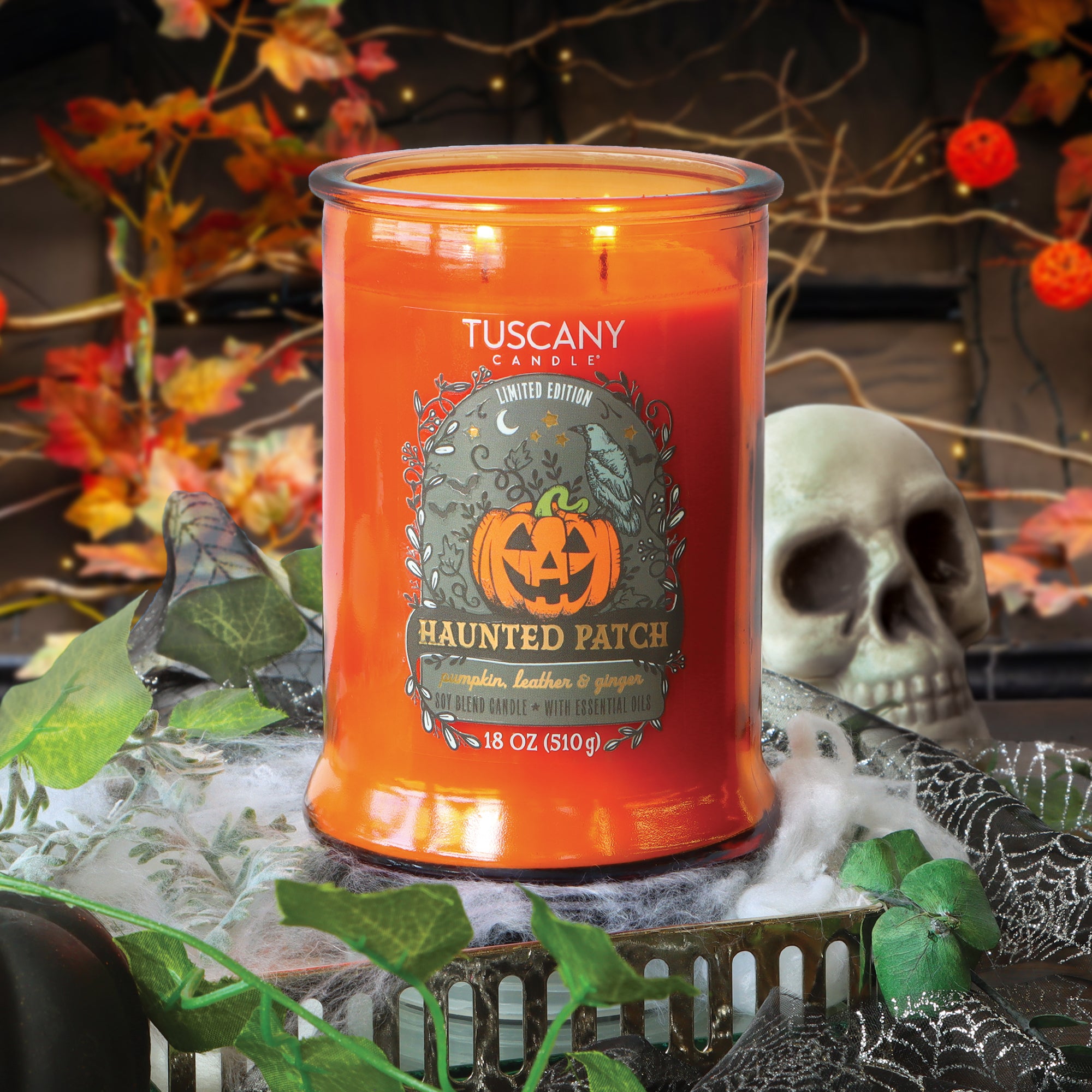 A Tuscany Candle® SEASONAL “Haunted Patch: Pumpkin, Leather & Ginger Scented Halloween Candle” (18 oz) rests on a table decorated with cobwebs, a skull, and autumn leaves in a festive Halloween scene.