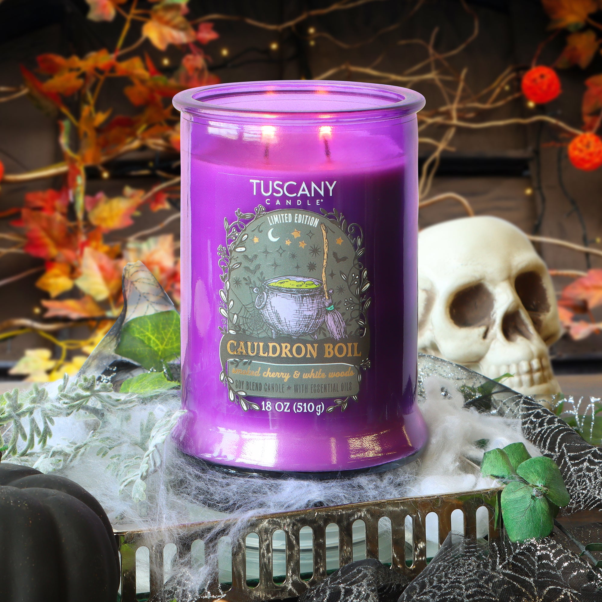 An 18 oz Tuscany Candle® SEASONAL “Cauldron Boil: Smoked Cherry & White Woods” scented Halloween candle sits on a tray surrounded by cobwebs, a skull, autumn leaves, and small pumpkins in the background.