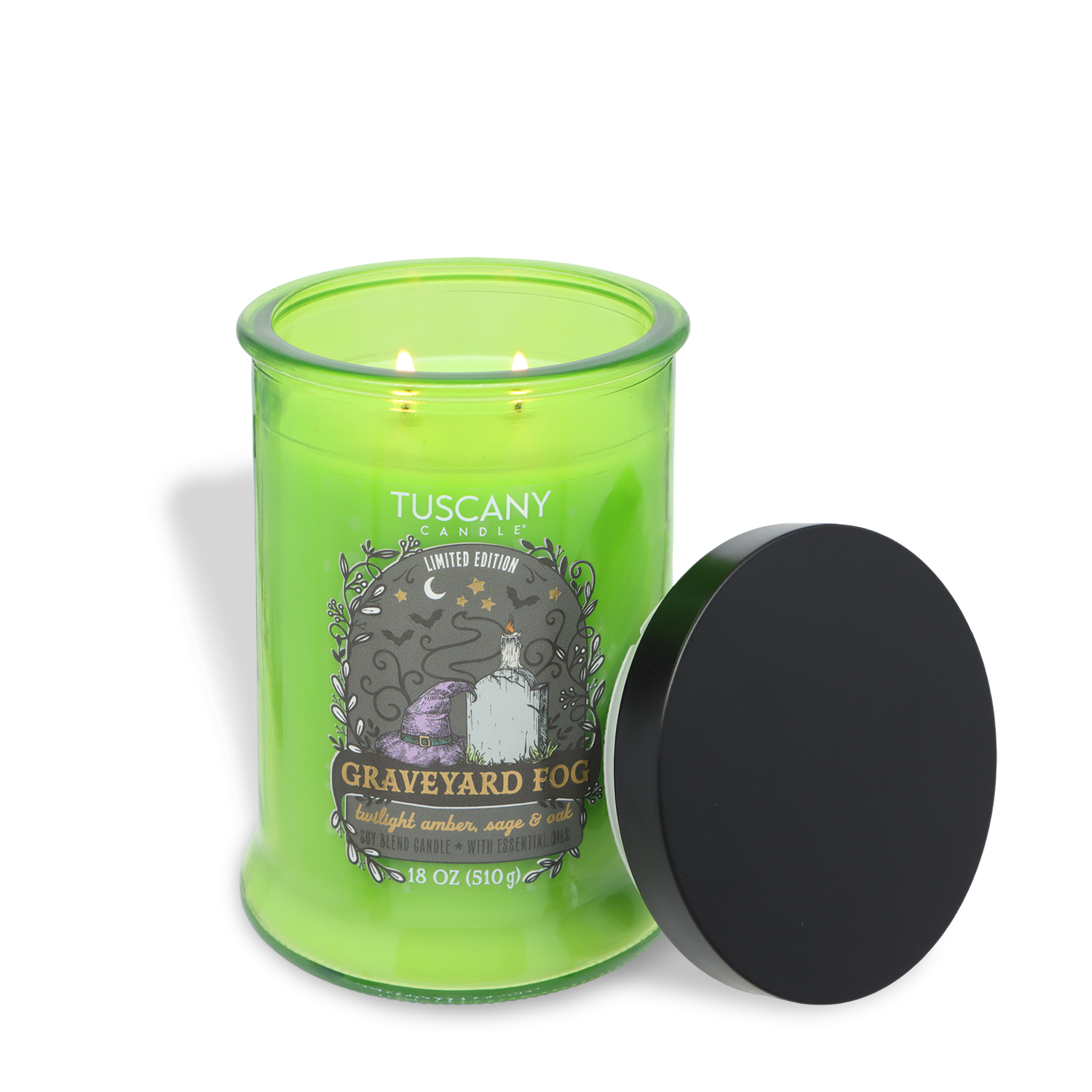 A green Tuscany Candle® SEASONAL jar labeled “Graveyard Fog: Twilight Amber, Sage & Oak” (18 oz) with two wicks burning and a black lid resting against the glass container.
