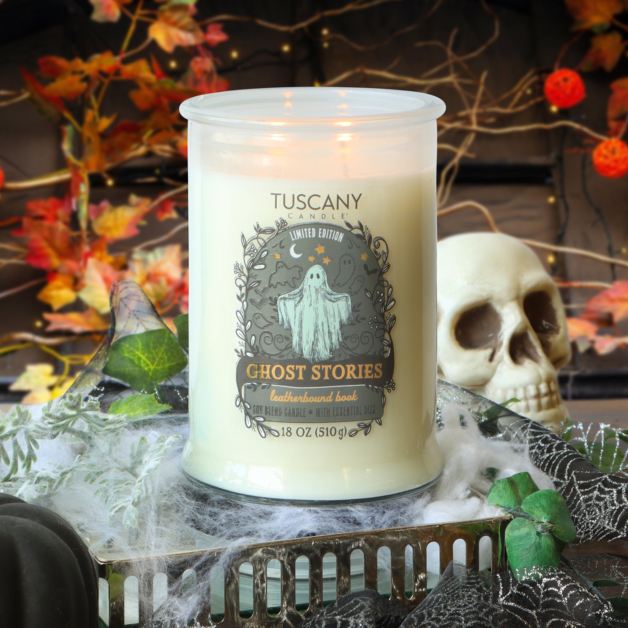 A lit Tuscany Candle® SEASONAL “Ghost Stories: Leatherbound Book Scented Halloween Candle” (18 oz) sits on a spiderweb-covered tray, surrounded by autumn leaves, a skull decoration, and Halloween props.