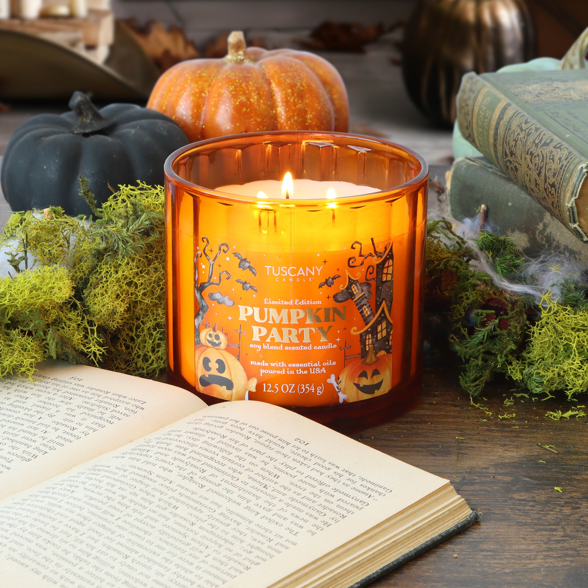 A Tuscany Candle® SEASONAL “Pumpkin Party: Clove & Tonka Scented Halloween Candle” (12.5 oz) glows on a table adorned with moss, pumpkins, an open book, and an old closed book in the background.