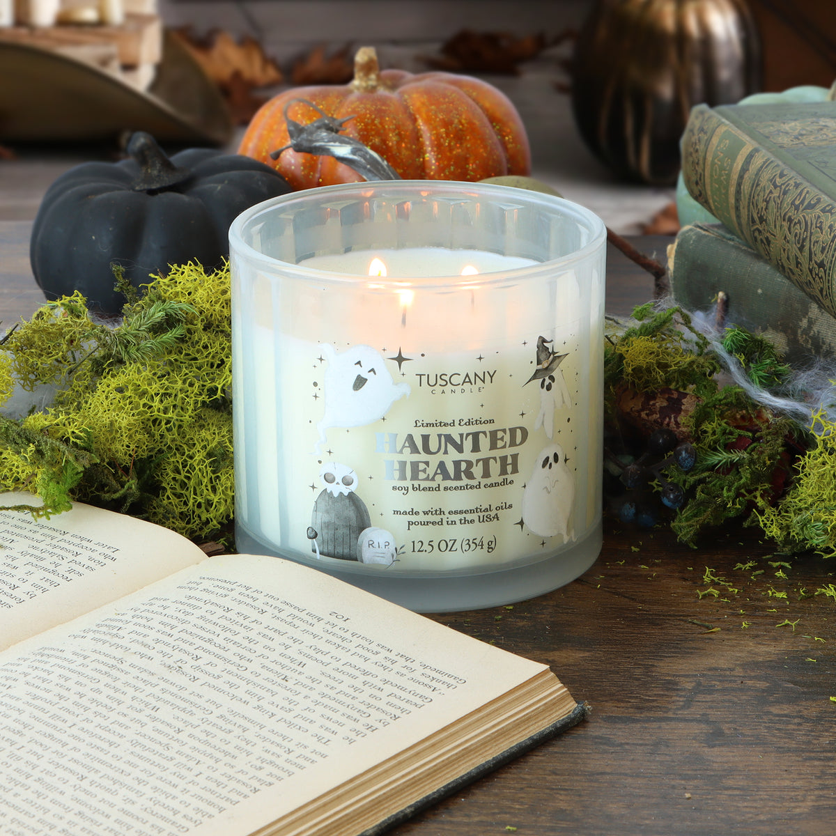 A lit Tuscany Candle® SEASONAL “Haunted Hearth: Apple & Citrus Zest Scented Halloween Candle” (12.5 oz) sits on a wooden table adorned with moss, books, pumpkins, and an open book nearby.