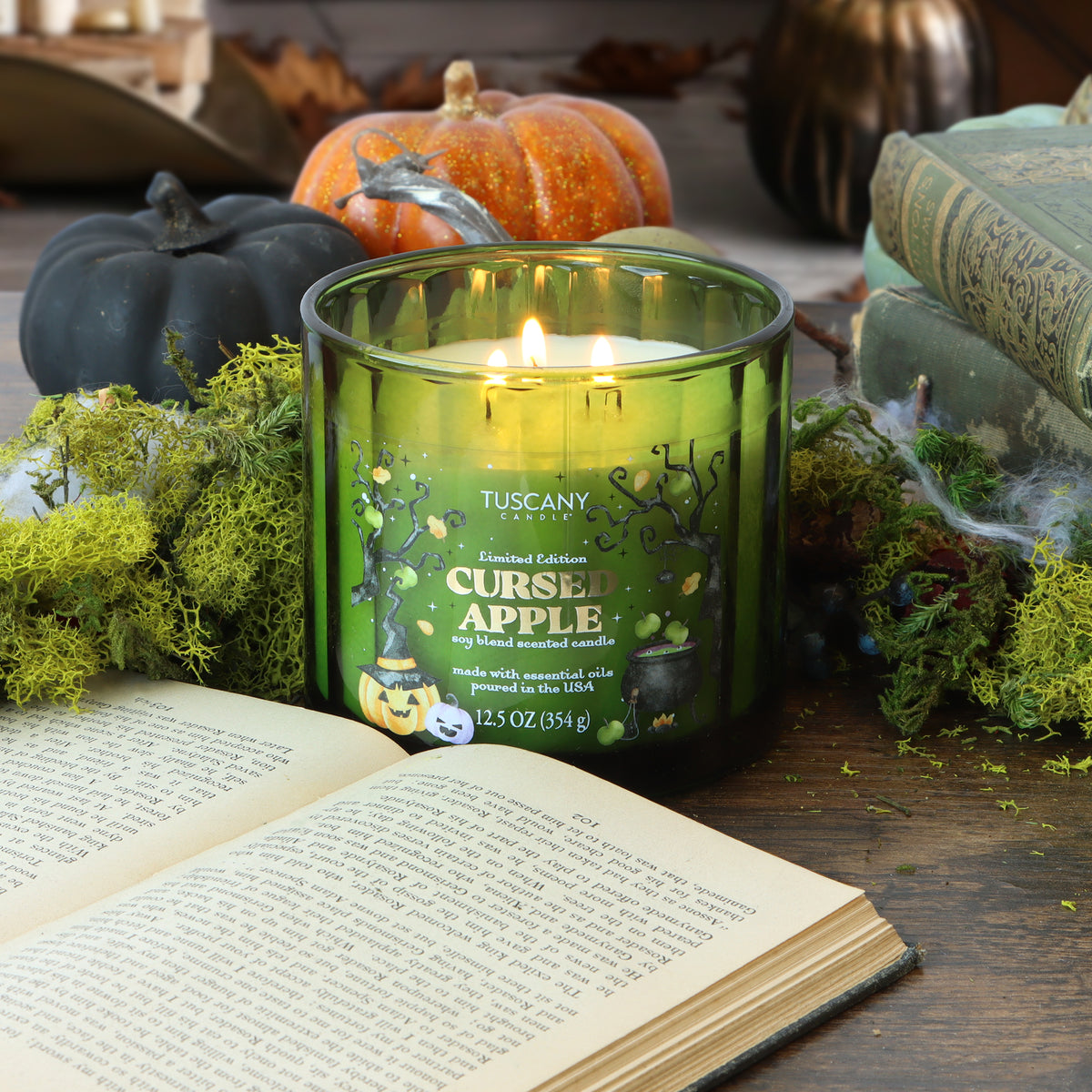 A lit “Cursed Apple: Nutty Caramel Apple Scented Halloween Candle” by Tuscany Candle® SEASONAL (12.5 oz) sits on a wooden table surrounded by moss, pumpkins, and an open book, creating a cozy autumn atmosphere.
