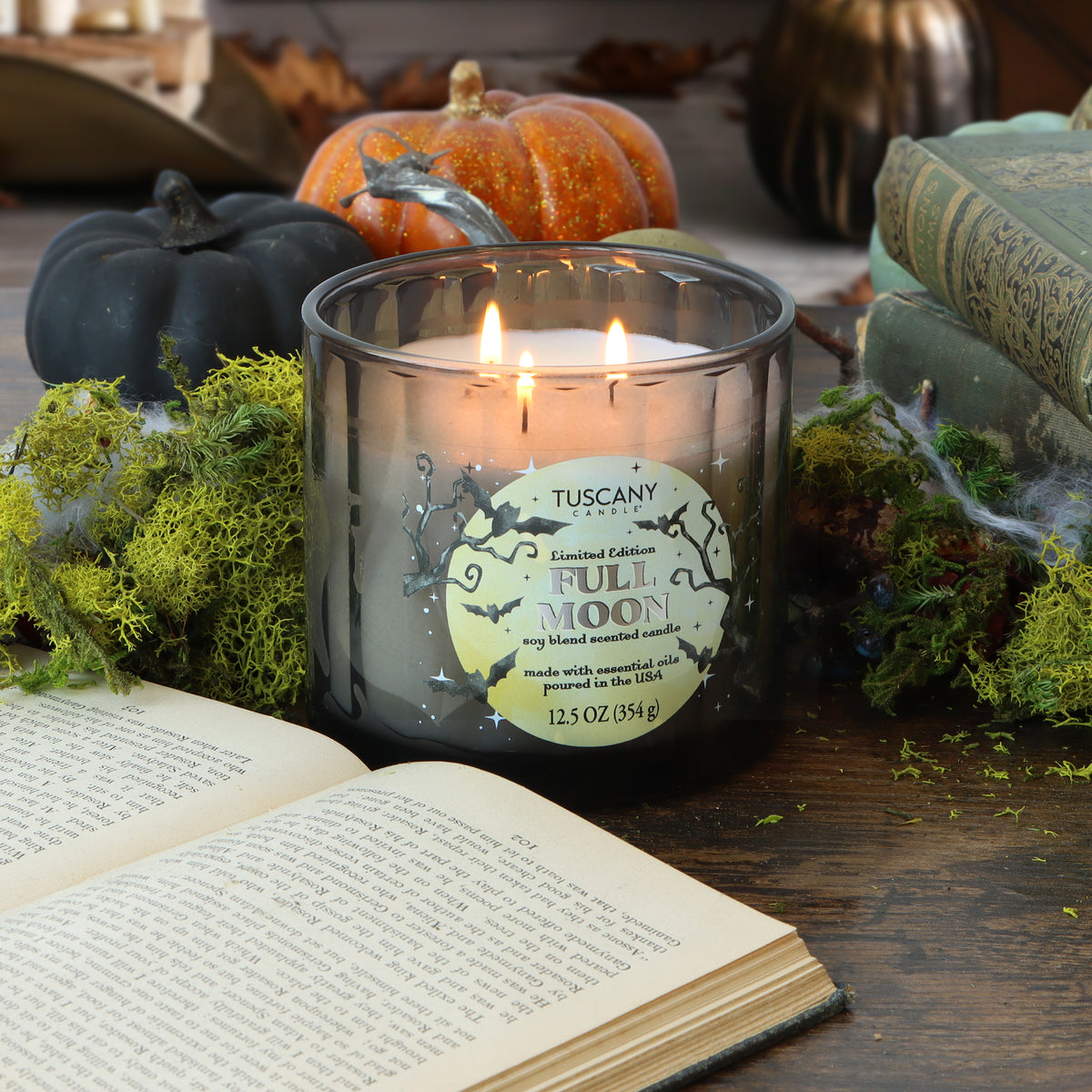 A Tuscany Candle® SEASONAL “Full Moon: Black Timber & Red Cherry Scented Halloween Candle” (12.5 oz) glows on a table adorned with moss, pumpkins, and books—an open book in the foreground—setting the scene for a cozy autumn atmosphere.