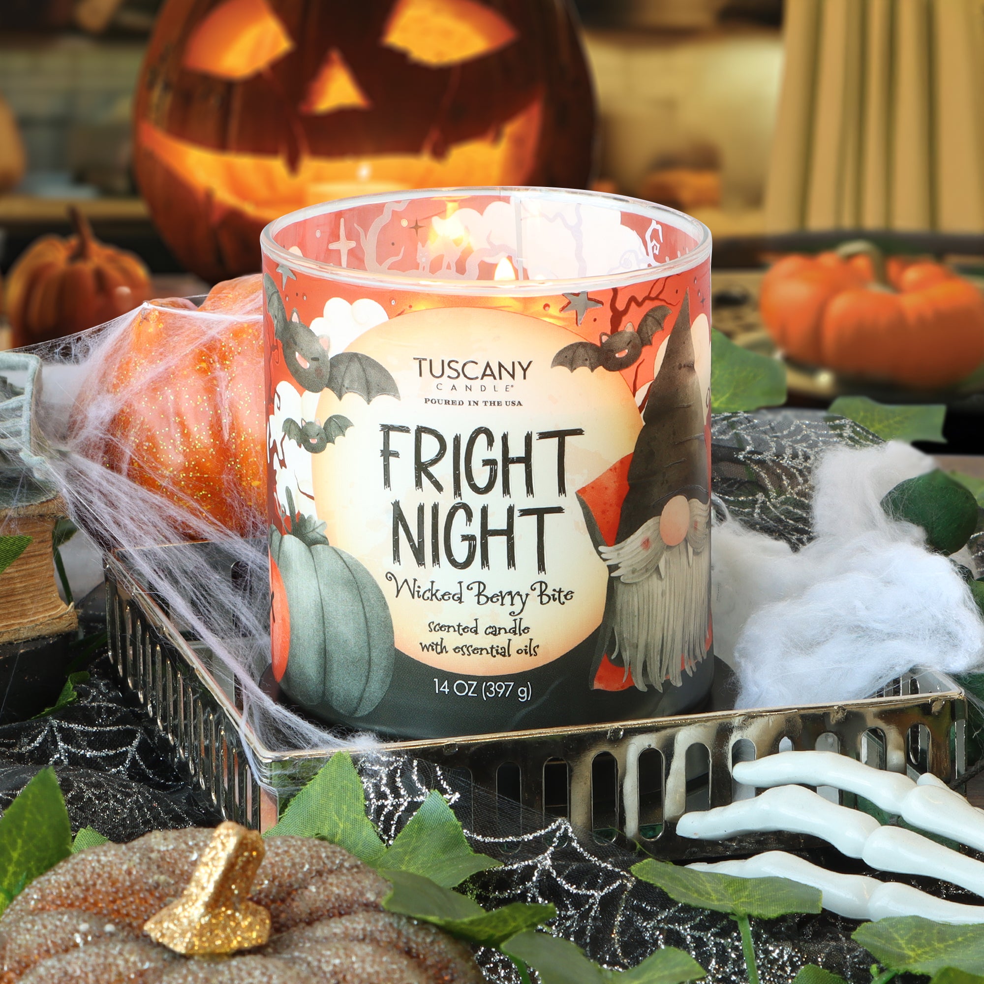 A Tuscany Candle® SEASONAL Fright Night: Wicked Berry Bite Scented Halloween Candle (14 oz) with festive Halloween-themed graphics sits on a tray surrounded by pumpkins, a spiderweb cloth, and spooky Halloween decorations.